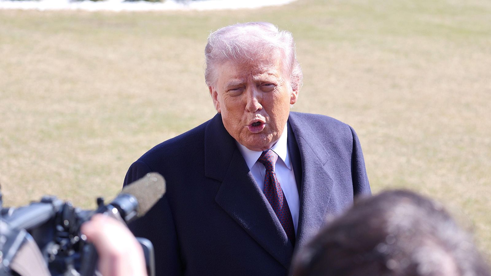 Donald Trump, con traje oscuro y corbata estampada, habla ante micrófonos de varios medios. Se aprecia un fondo con césped y parte de un edificio, posiblemente en un entorno oficial.