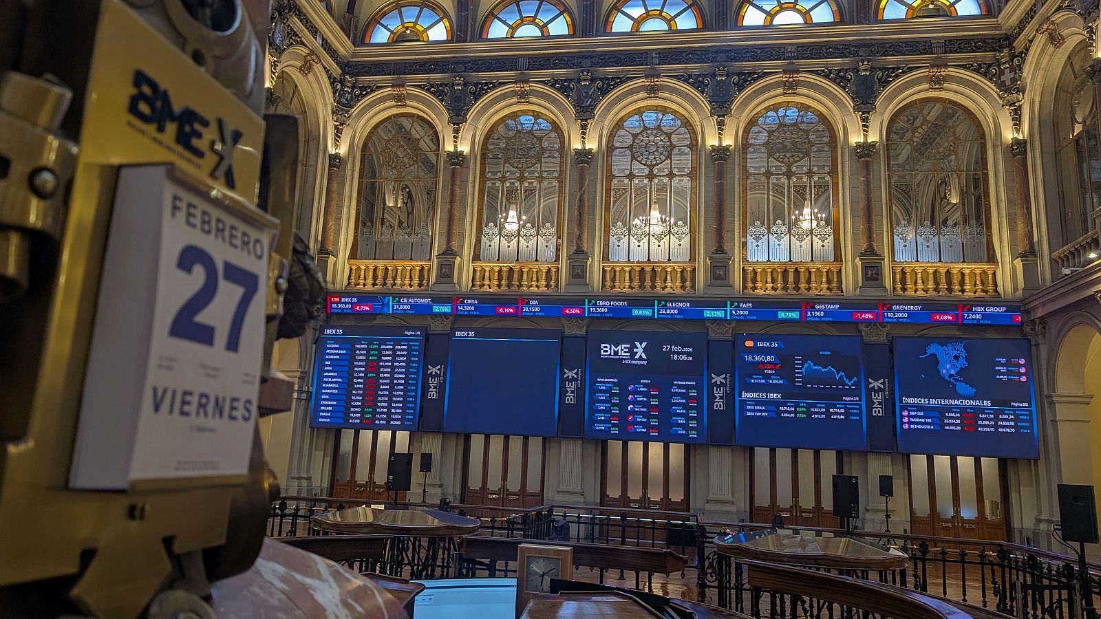 En el interior de un edificio con arquitectura clásica, se observa un calendario que marca el 27 de febrero, un viernes. Al fondo, varias pantallas muestran información financiera y gráficos bursátiles.