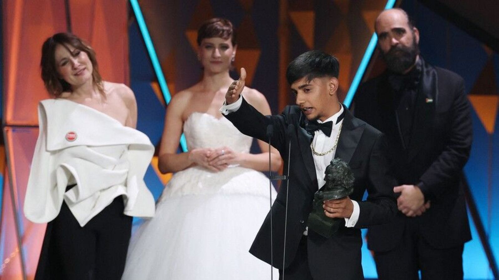 Antonio Fernández Gabarre, con un esmoquin negro y pajarita, celebra su premio sosteniendo la estatuilla y agradeciendo. A su lado, tres personas observan la escena, una con un conjunto blanco y negro, otra con un vestido blanco y un hombre con traje negro.