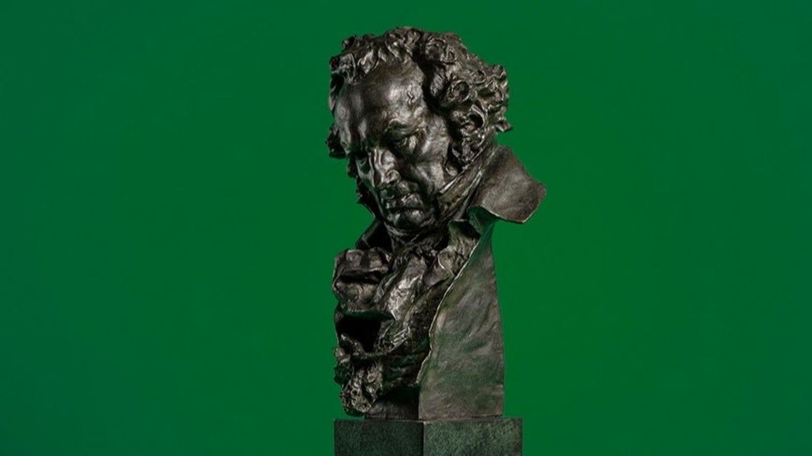 El primer plano muestra el premio Goya, una escultura de bronce que representa el busto de un hombre sobre un pedestal, con un fondo verde.