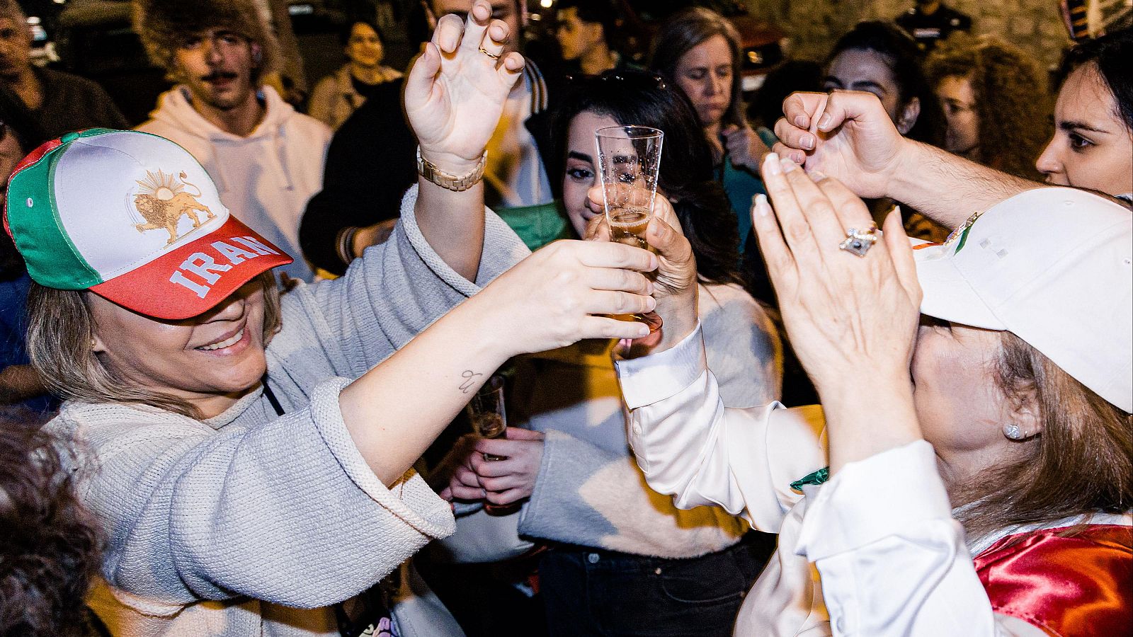 Un grupo celebra con brindis, destacando una persona con una gorra con la bandera iraní y la palabra "IRAN". Se observan más personas al fondo, algunas mirando a la cámara.
