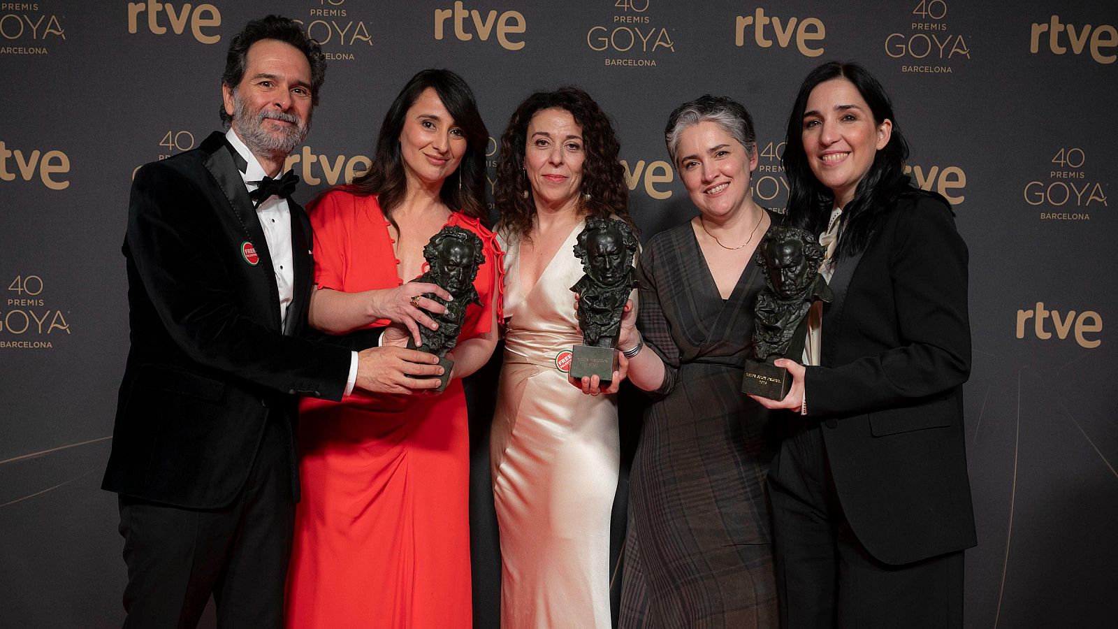 Cinco personas, incluyendo un hombre en esmoquin y varias mujeres con vestidos largos, celebran con estatuillas de los Premios Goya. El fondo oscuro muestra logotipos y la inscripción de la edición en Barcelona.
