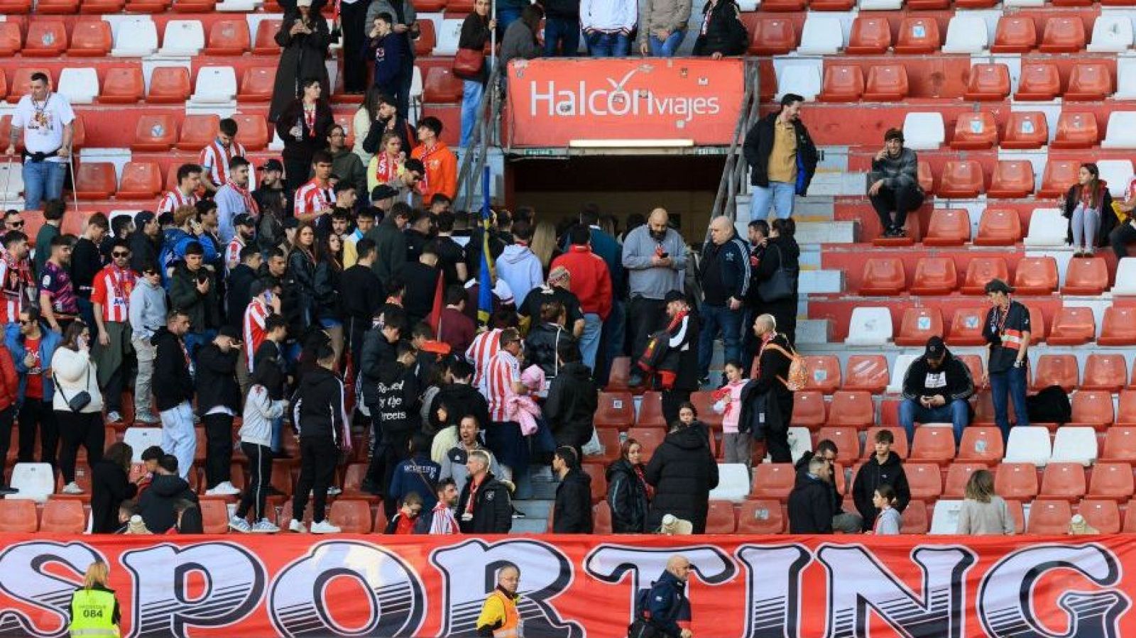 Aficionados del Sporting de Gijón abandonan el estadio tras un evento deportivo. Se aprecia una pancarta con el nombre del equipo y un anuncio publicitario, con las gradas parcialmente vacías y personal de seguridad presente.