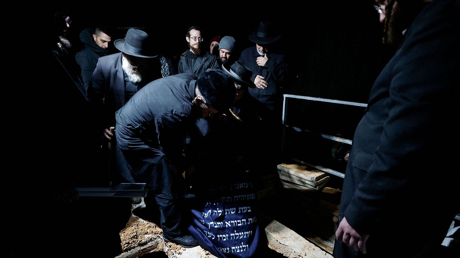 En un cementerio, un grupo de personas se reúne alrededor de una tumba. La escena, con una iluminación tenue, sugiere un momento de luto y recogimiento. Se observan hombres vestidos con trajes oscuros.