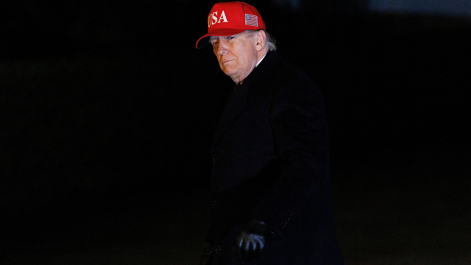 Donald Trump, con una gorra roja con las letras "USA" y la bandera estadounidense, y un abrigo oscuro, posa en un ambiente de poca luz, posiblemente de noche.