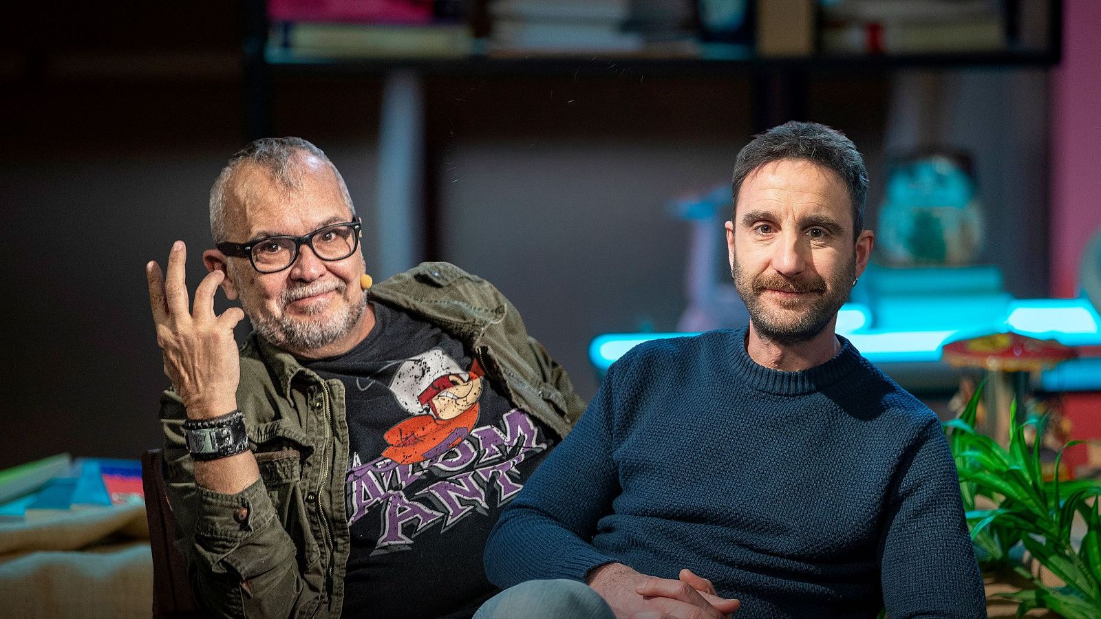 Dos hombres posan en un estudio; uno con gafas y camiseta con un dibujo, sonríe y levanta la mano, mientras el otro, con jersey azul y barba, mira a la cámara con serenidad. El fondo incluye estanterías y el título "Mi persona favorita".