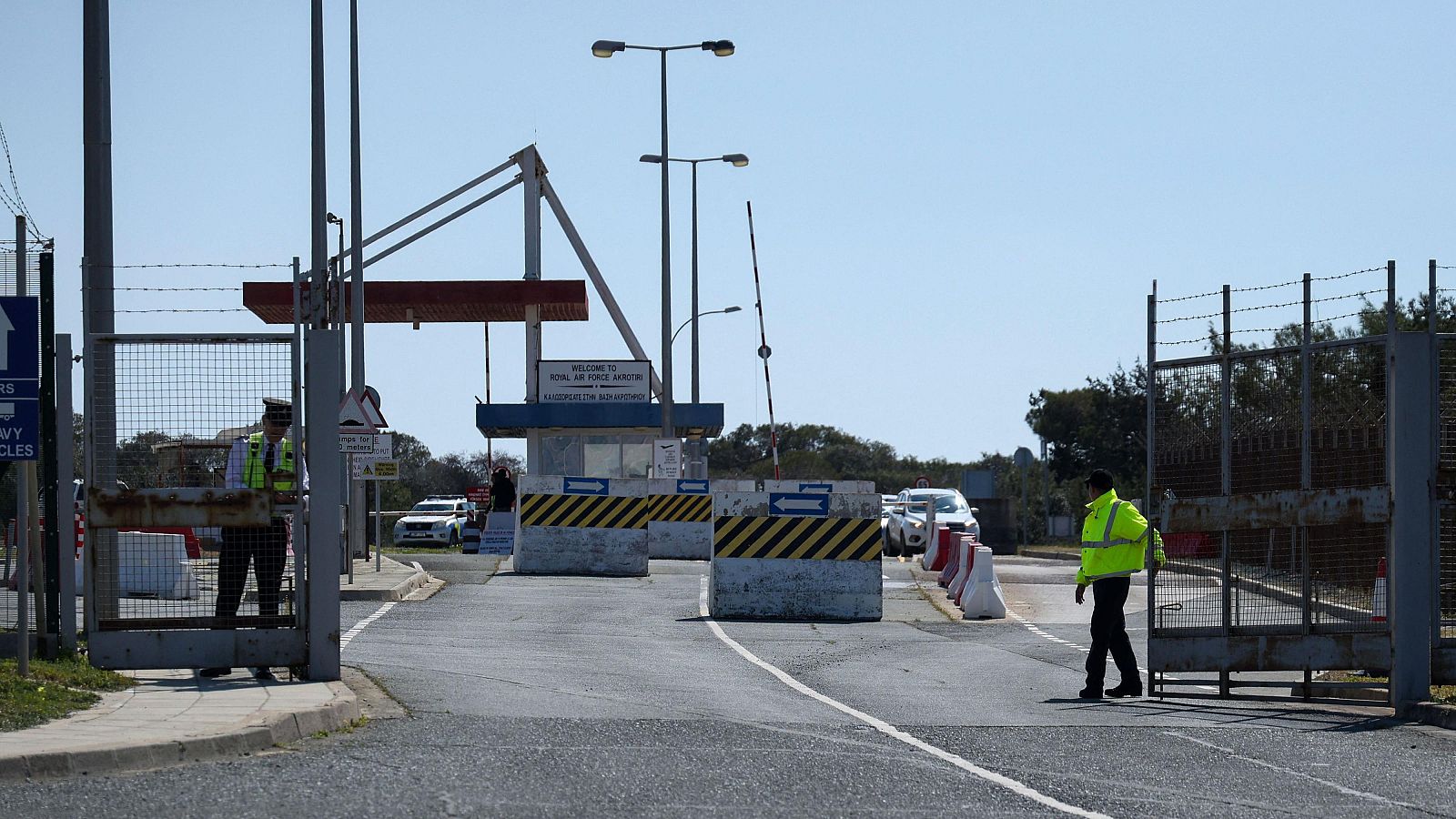 En la entrada de una base militar, una carretera asfaltada conduce a puertas metálicas con alambre de púas. Dos guardias de seguridad uniformados vigilan la entrada, mientras una caseta anuncia la base con un letrero.