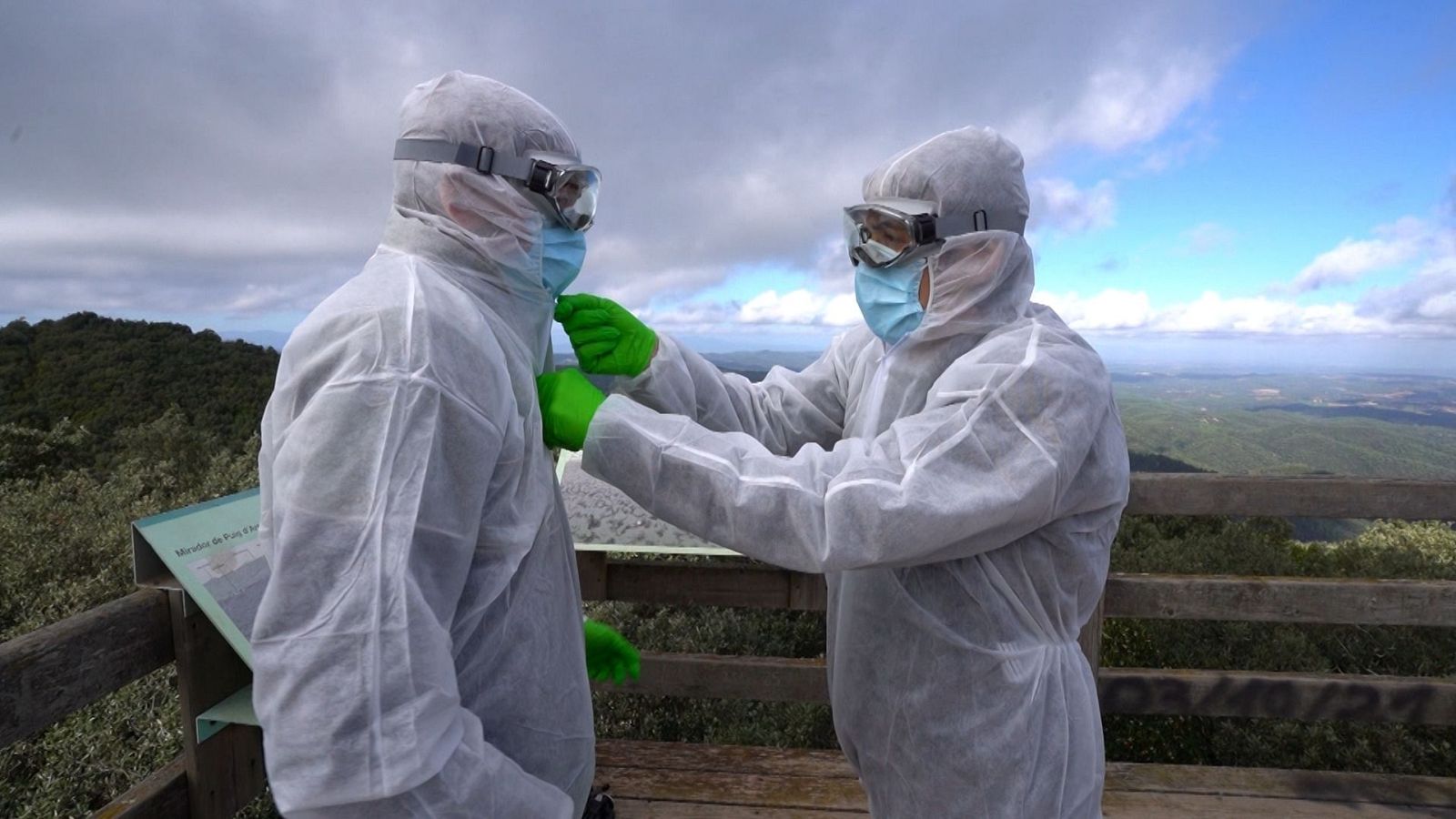 Dues persones amb vestits de protecció blancs, mascaretes i guants, en una plataforma de fusta amb barana, amb un paisatge muntanyós de fons.