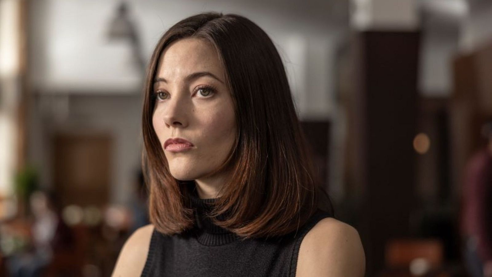 Una mujer de piel clara, con cabello castaño oscuro y ojos marrones, posa con un jersey negro de cuello alto y labios rojos, en un ambiente interior con iluminación tenue.