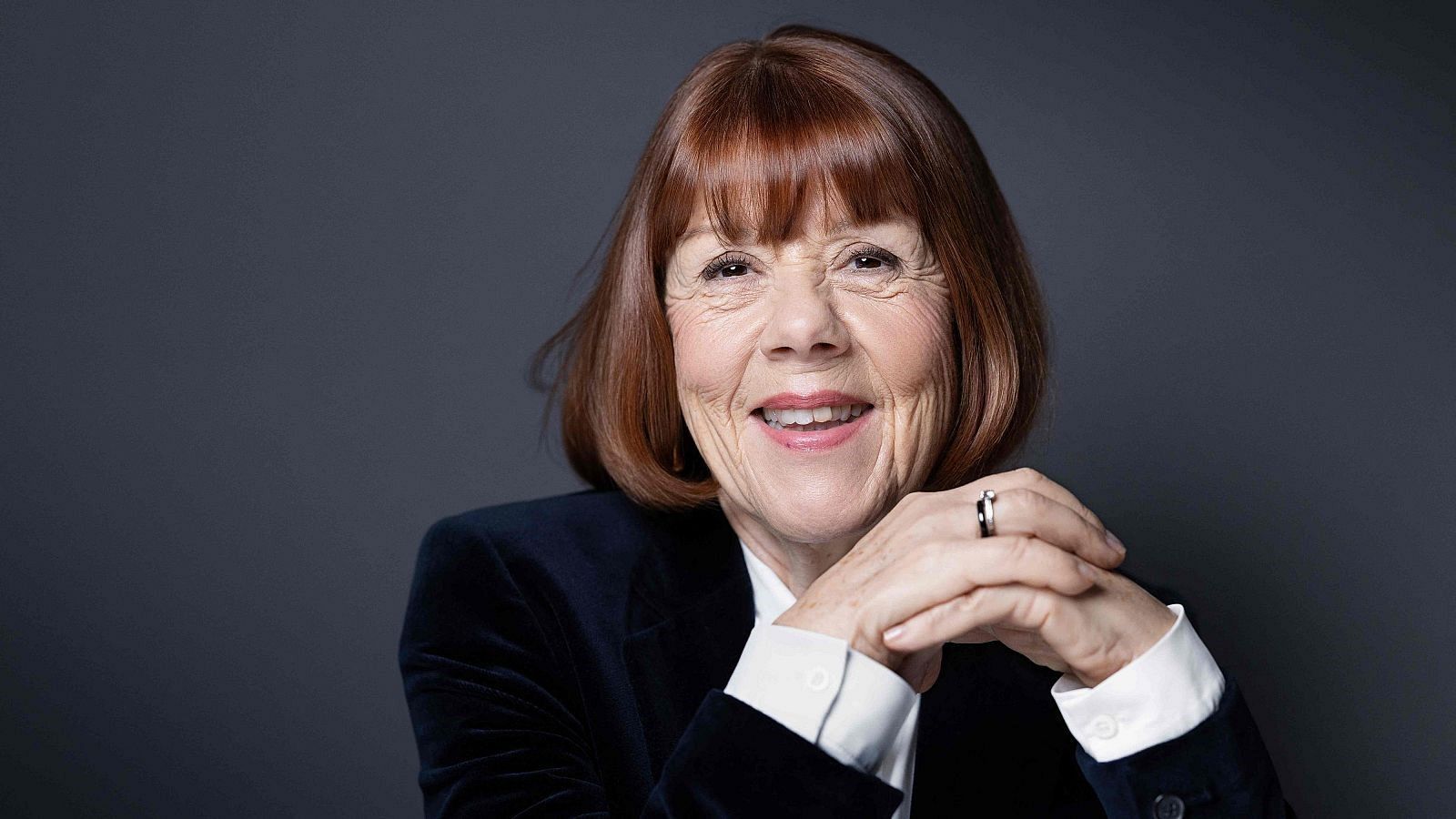 Una mujer mayor, con cabello castaño rojizo y flequillo, sonríe a la cámara. Viste una chaqueta oscura sobre una camisa clara y lleva un anillo en la mano izquierda, con las manos entrelazadas.