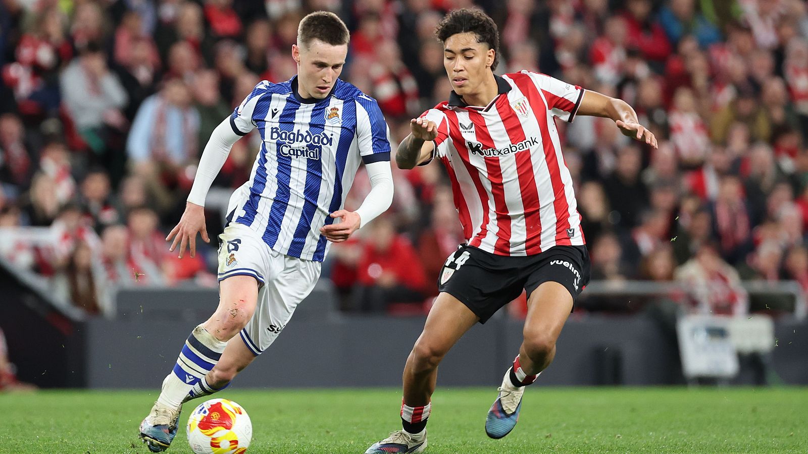 Un jugador de la Real Sociedad, con camiseta a rayas, conduce el balón ante la presión de un jugador del Athletic, también con camiseta a rayas, en un partido de fútbol con público.
