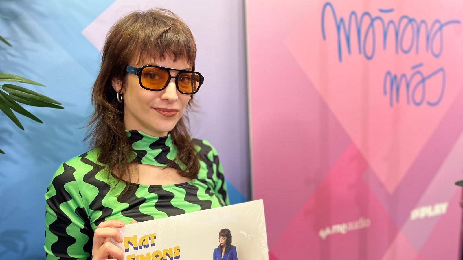 Una mujer con gafas de sol y una blusa estampada sostiene un disco frente a una pared con diseños geométricos y la frase "Mañana más".