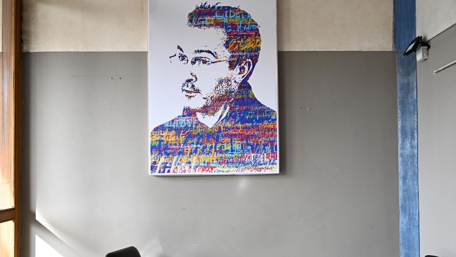 Un retrato colorido de un hombre con gafas y barba, realizado con trazos y palabras de diferentes colores, cuelga de una pared gris y blanca.