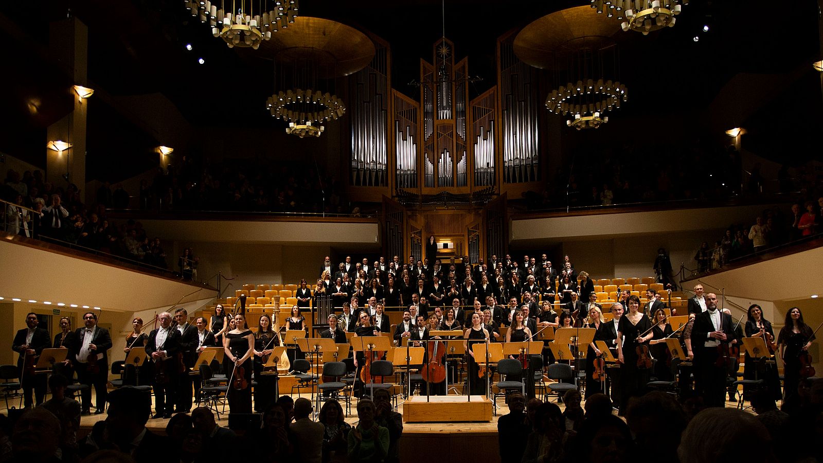 Una orquesta y un coro, vestidos formalmente, actúan en un escenario frente a un público en un auditorio. Se distinguen instrumentos musicales y un gran órgano al fondo, con el público atento a la presentación.