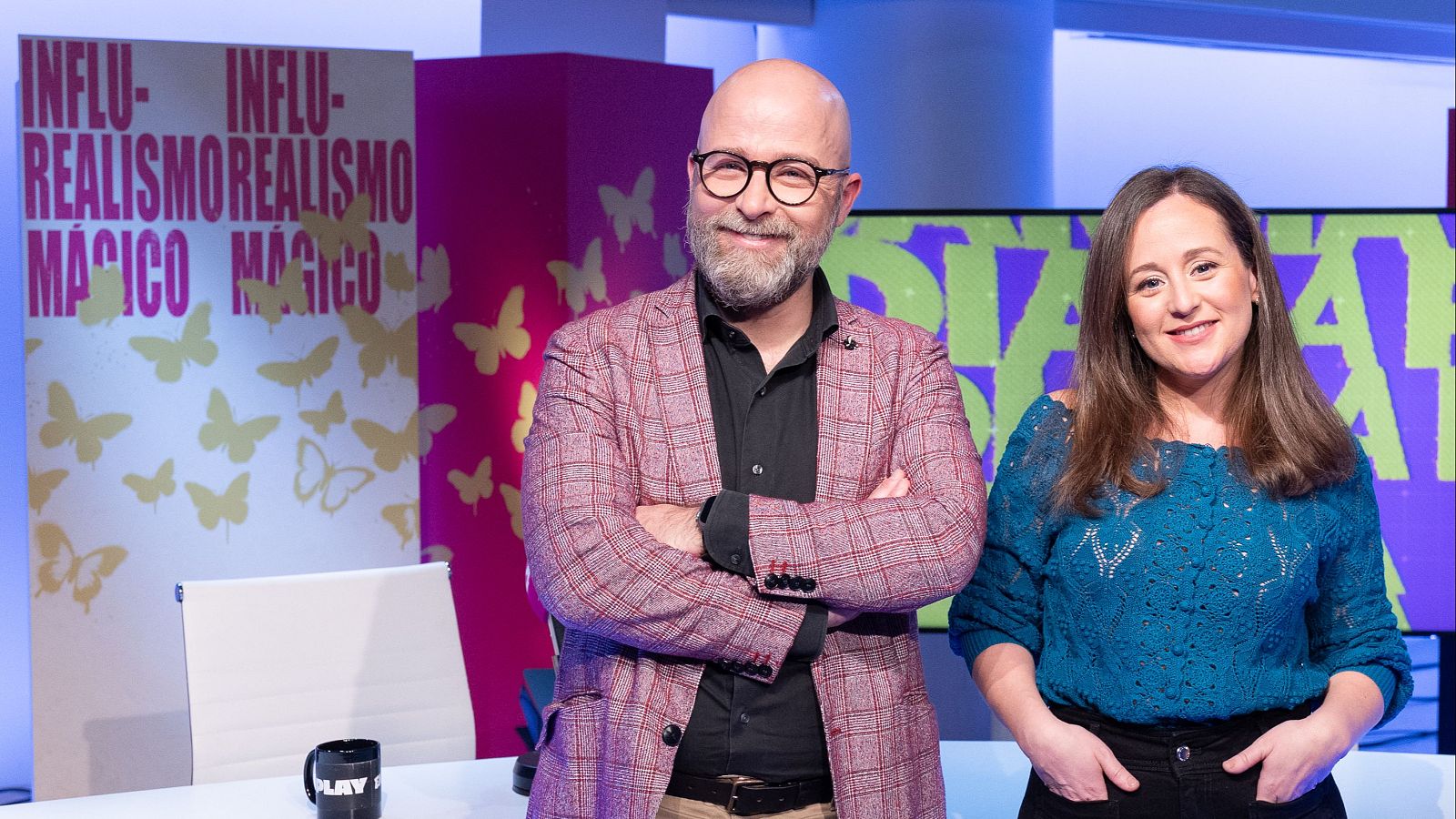 Dos personas sonríen en un estudio de radio. Un hombre con gafas y chaqueta de cuadros, y una mujer con jersey azul, posan frente a carteles con un diseño repetitivo y una taza con la palabra 'PLAY'.
