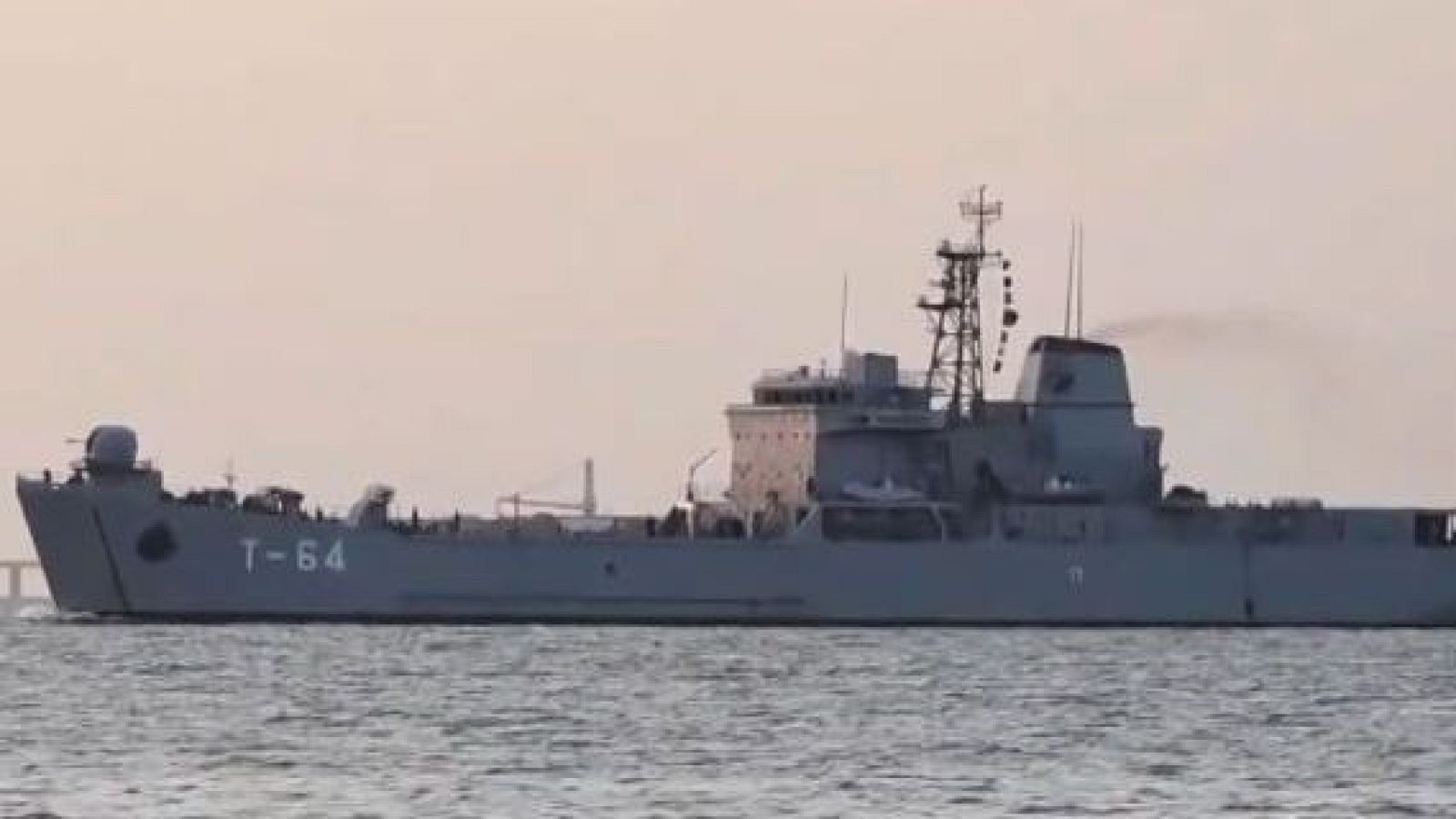 Un barco gris, posiblemente un petrolero, surca las aguas con la inscripción "T-64" visible en su costado, acompañado de antenas, una chimenea y un puente al fondo, bajo un cielo despejado.