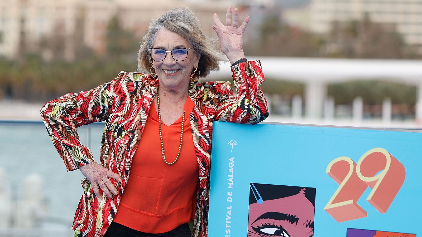 Carmen Maura, con gafas y cabello canoso, sonríe y saluda. Viste una chaqueta estampada, una blusa naranja y un collar dorado, apoyada en un cartel con el número 29.