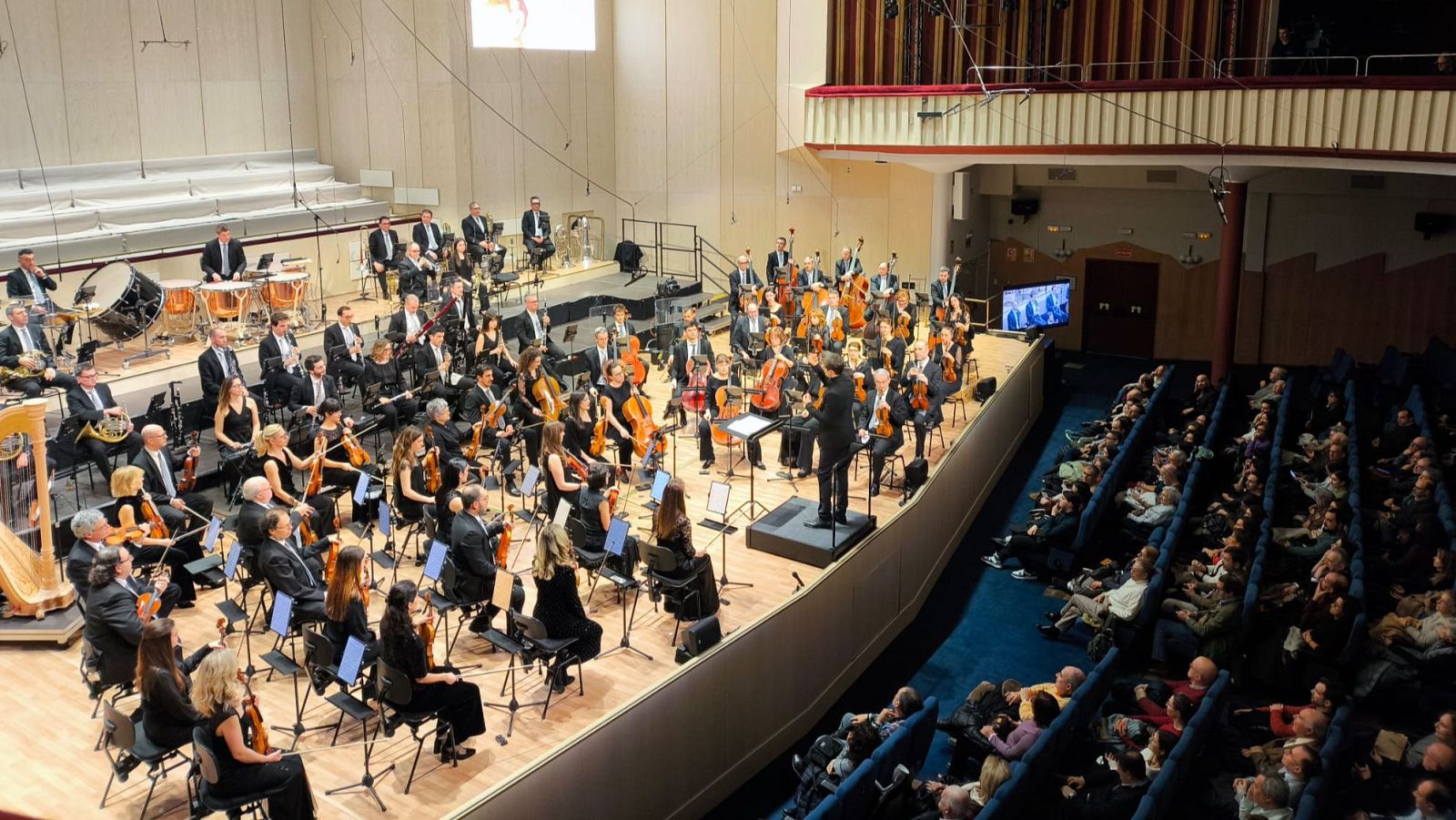 Una orquesta con numerosos músicos, incluyendo instrumentistas de cuerda y viento, se presenta en un escenario. Un director dirige la interpretación, mientras el público observa el concierto.