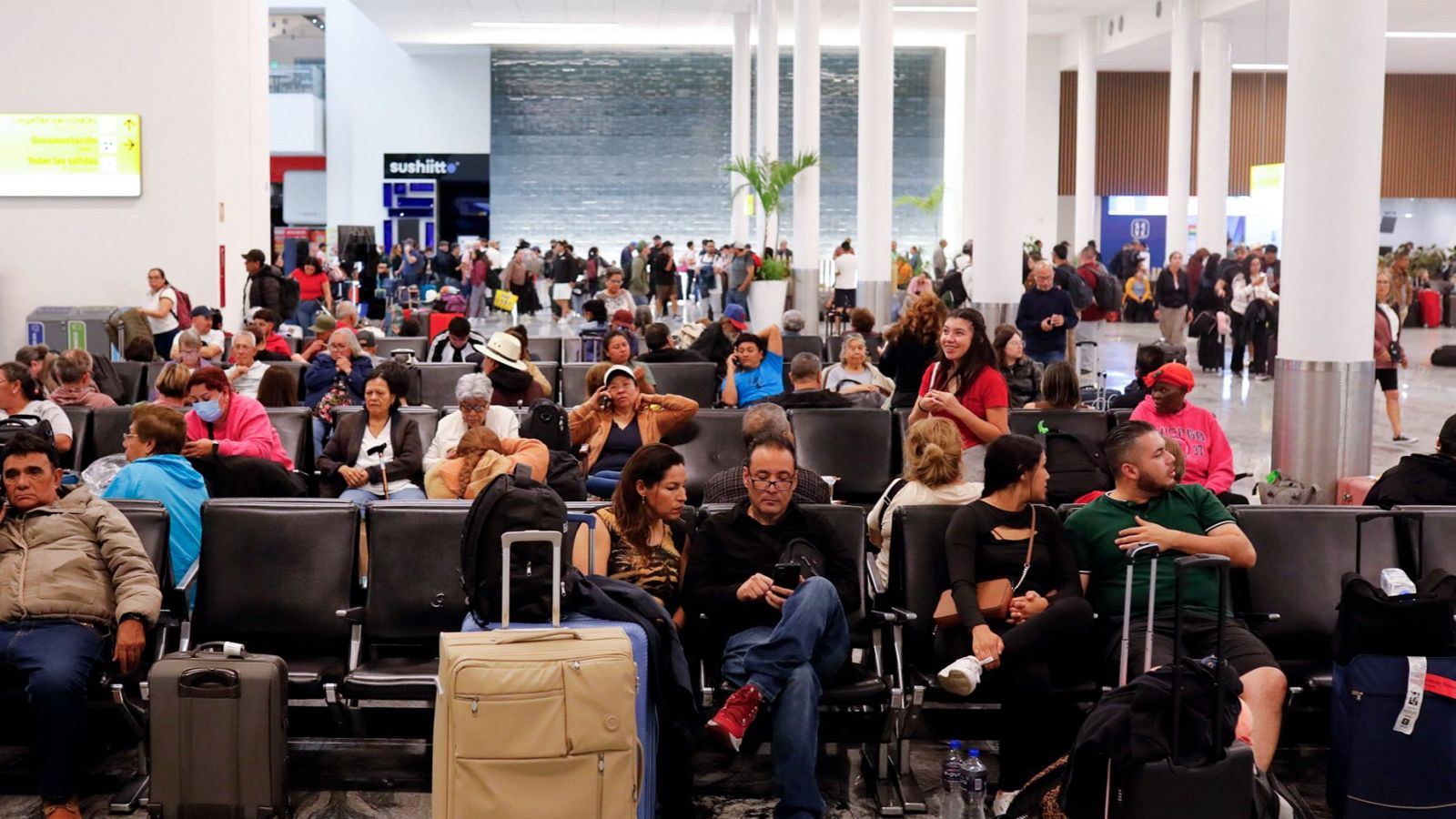 En una concurrida sala de espera de aeropuerto, se ven personas esperando, algunas sentadas y otras de pie, con equipaje. Una pantalla con información y un letrero con una palabra en japonés son visibles.