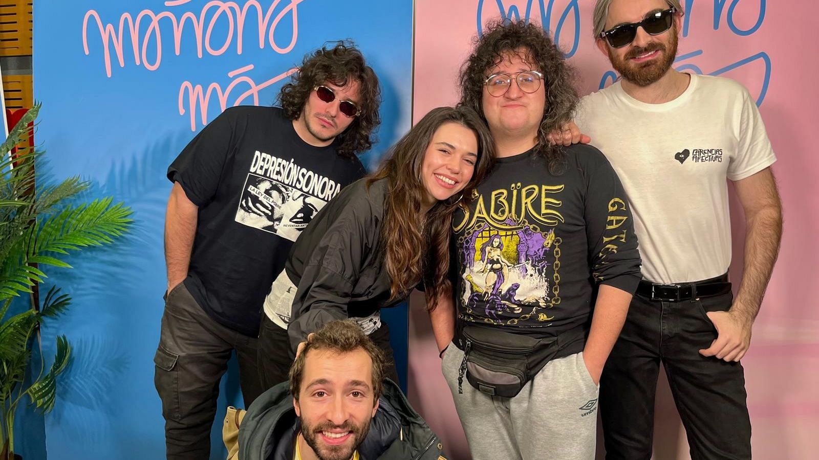 Cinco personas posan frente a un fondo dividido en azul y rosa, con la frase "Mañana más". Visten camisetas con estampados como "DEPRESIÓN SONORA" y "SABIRE GATEWAY", además de gafas de sol y otras prendas.