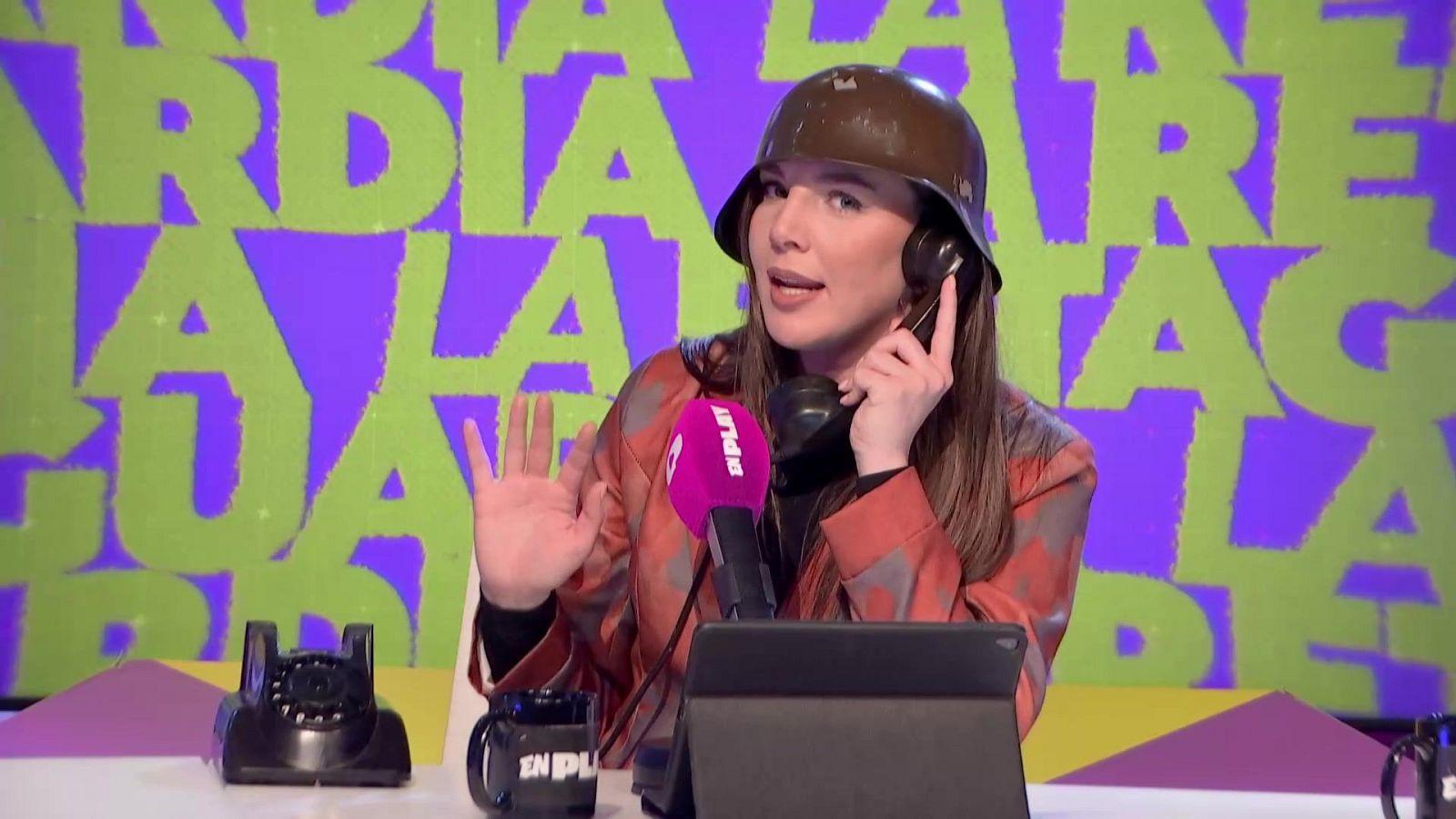 Una mujer con chaqueta naranja y marrón y casco militar sostiene un auricular y levanta la mano. Frente a ella, un micrófono rosa con el logo "en Play", un teléfono antiguo, una taza y una tablet.