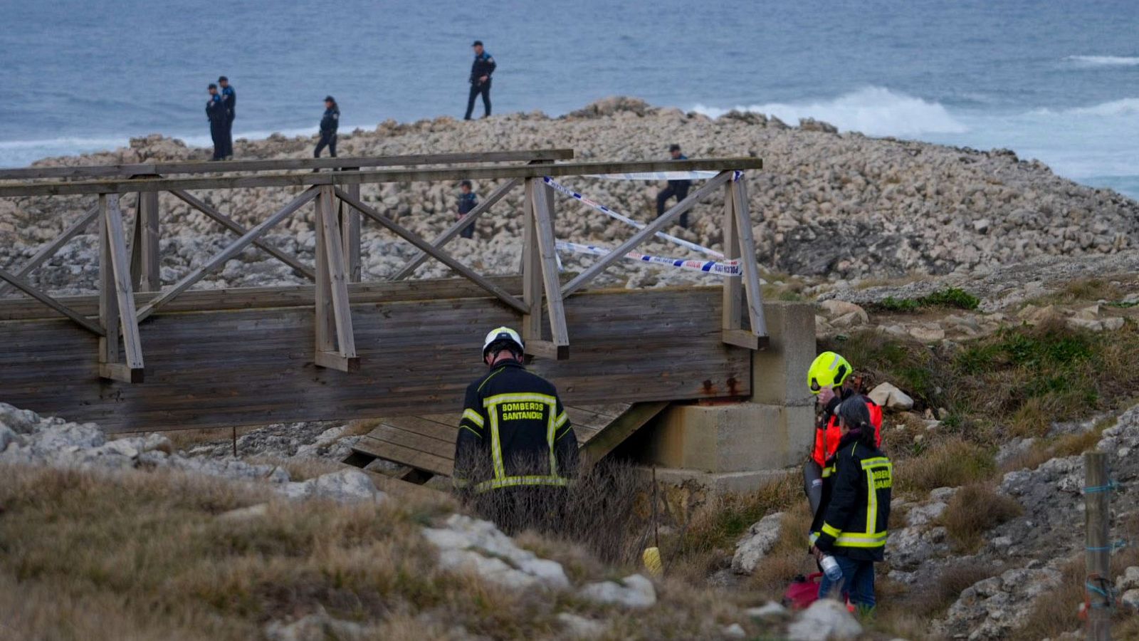 En la playa rocosa, un grupo de personas uniformadas, posiblemente bomberos, policías y personal sanitario, participan en una operación. Se observa una camilla en la escena, con el mar de fondo.