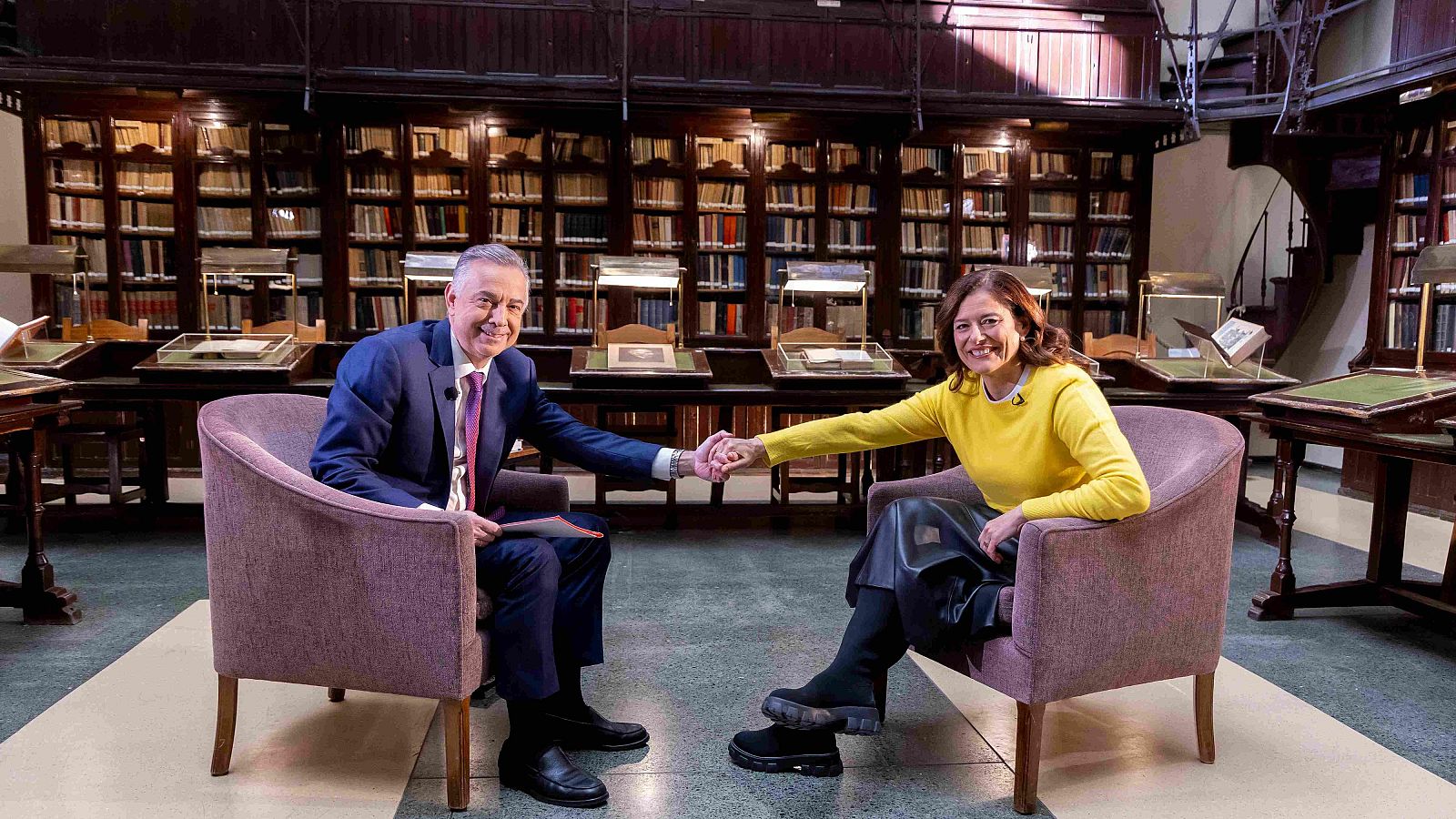 Dos personas, sonrientes, se dan la mano en una biblioteca. Una de ellas viste un jersey amarillo, falda y botas negras; la otra, un traje azul con corbata rosa.