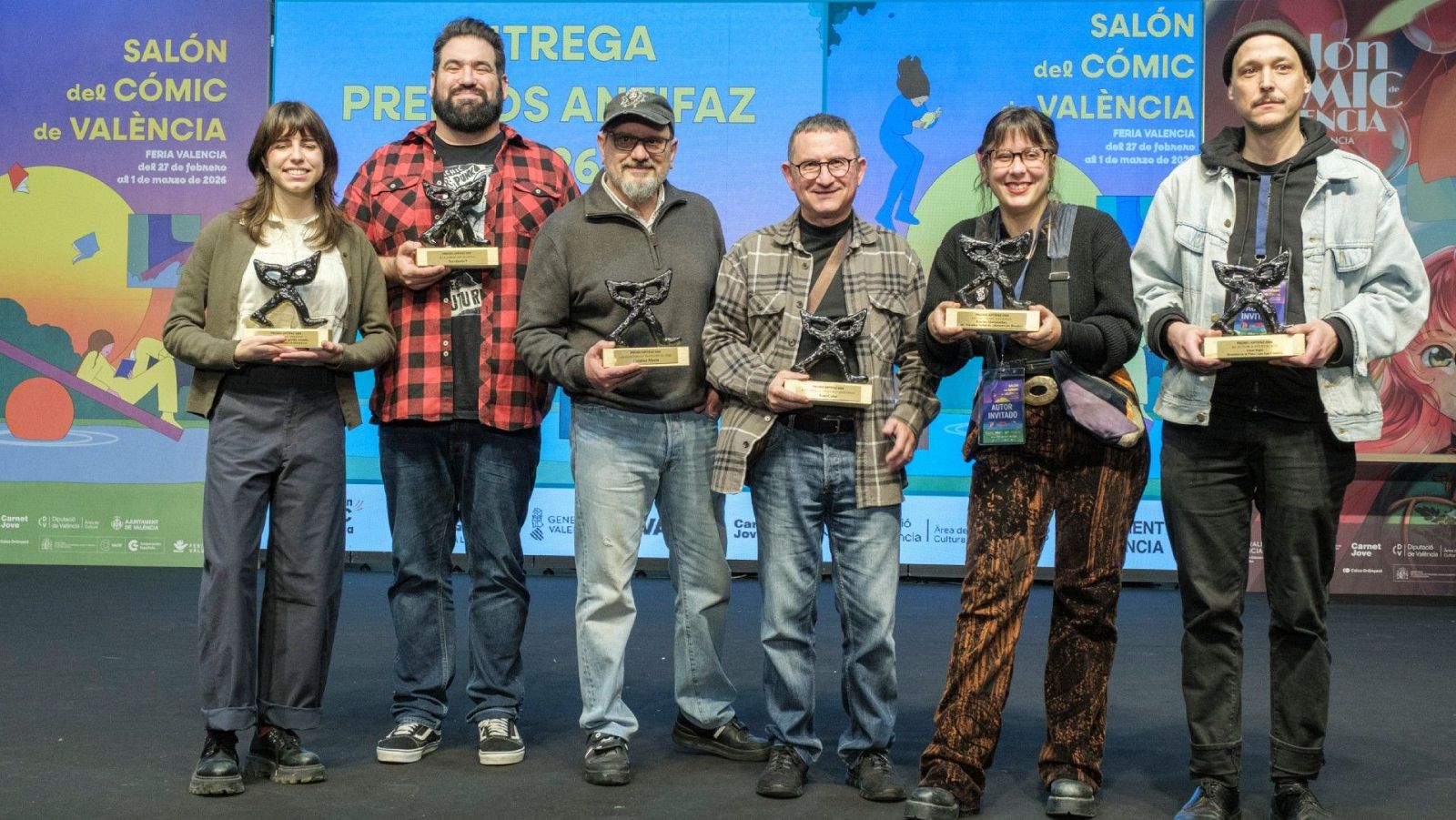 Seis personas posan en un escenario, cada una sosteniendo un premio con forma de máscara. El evento, celebrado en un salón, contó con la presencia de individuos de diferentes edades y vestimentas, destacando la diversidad de los galardonados.
