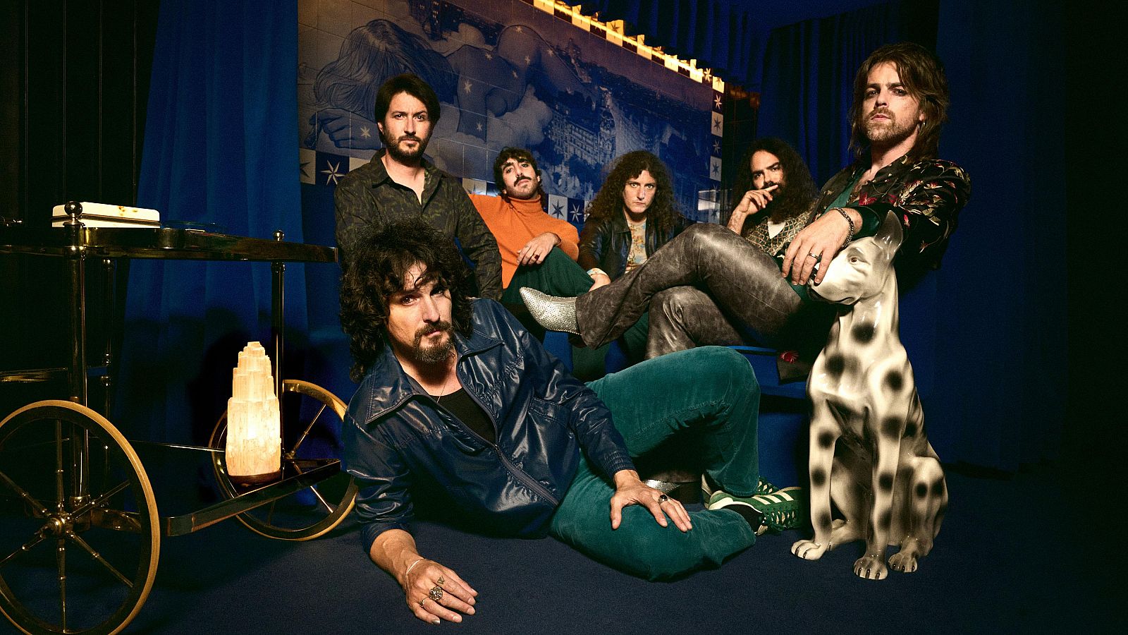 Fotografía de los seis integrantes de una banda musical, con vestimenta de estilo retro y psicodélico. Se observa un carrito de bar dorado con una lámpara de sal y una escultura de un perro dálmata, en un ambiente con iluminación cálida y tenue.