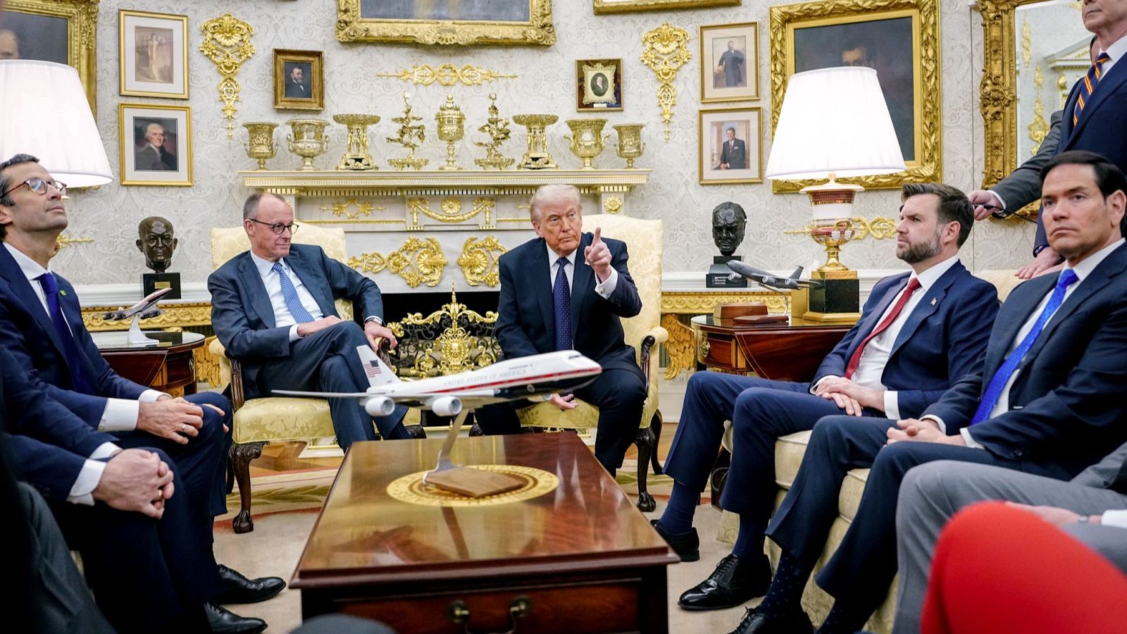 En un despacho ricamente decorado, dos hombres vestidos con trajes, uno de ellos señalando, parecen estar en una reunión. El entorno incluye paneles de madera, cuadros, molduras doradas y una maqueta de avión.