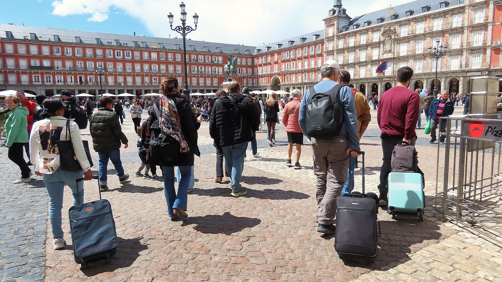 Turistas con maletas llenan la Plaza Mayor de Madrid en un día soleado, reflejando el récord turístico de España. Se aprecia la arquitectura de la plaza y parte de un cartel.