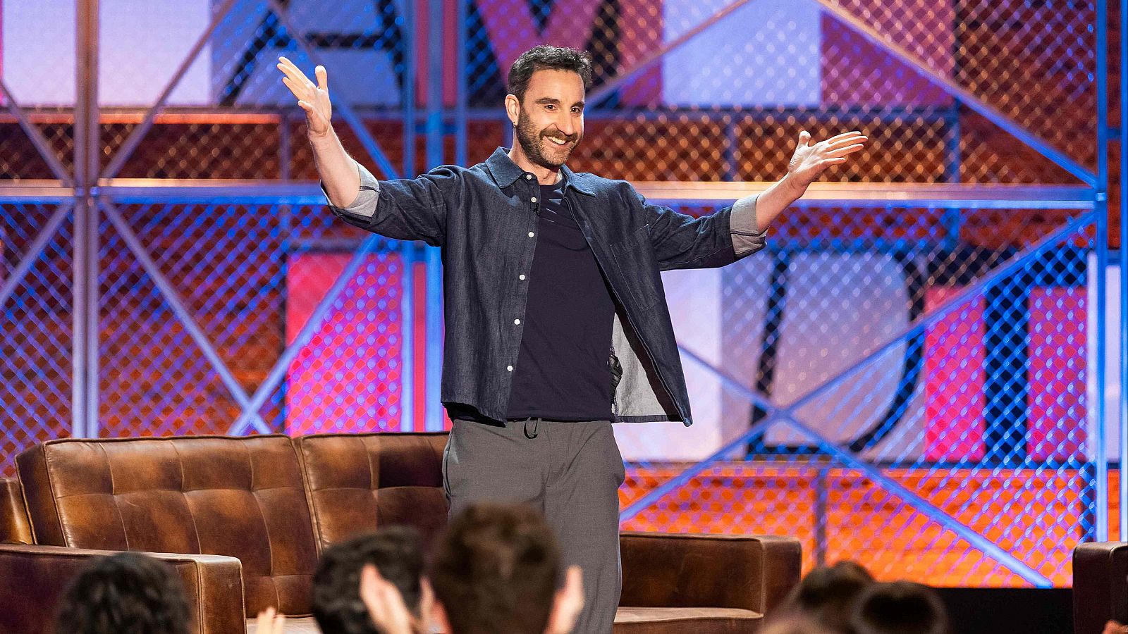 Dani Rovira, con una sonrisa y los brazos abiertos, posa en un escenario. Viste de manera informal, con una chaqueta vaquera y pantalones grises, frente a una estructura metálica con diseño geométrico. Se observa parte del público y un sofá.