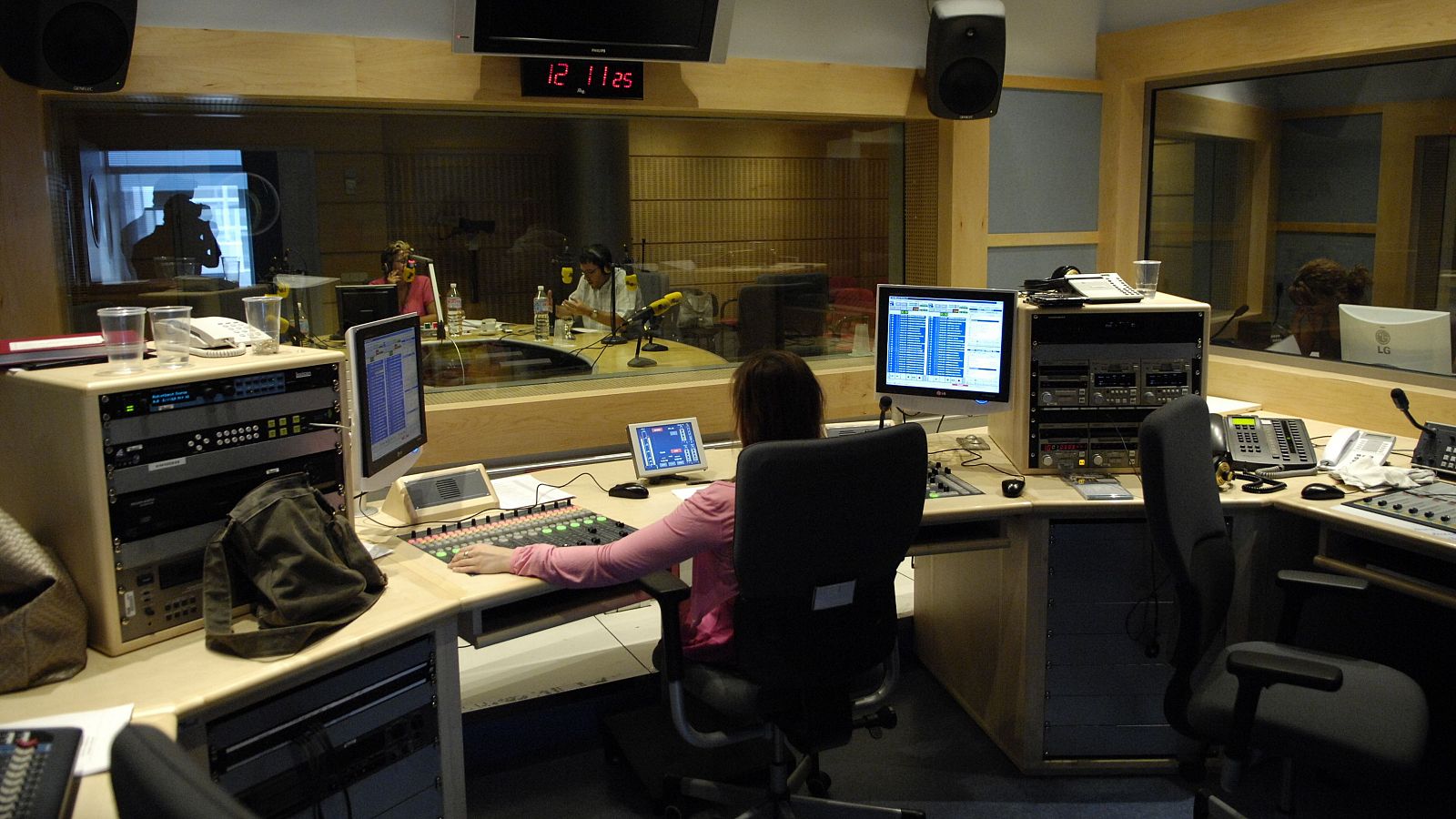 En un estudio de radio, una técnica opera una consola de sonido mientras una directora se prepara para la emisión. El ambiente es profesional y ordenado, con equipos de comunicación y un reloj digital marcando la hora.