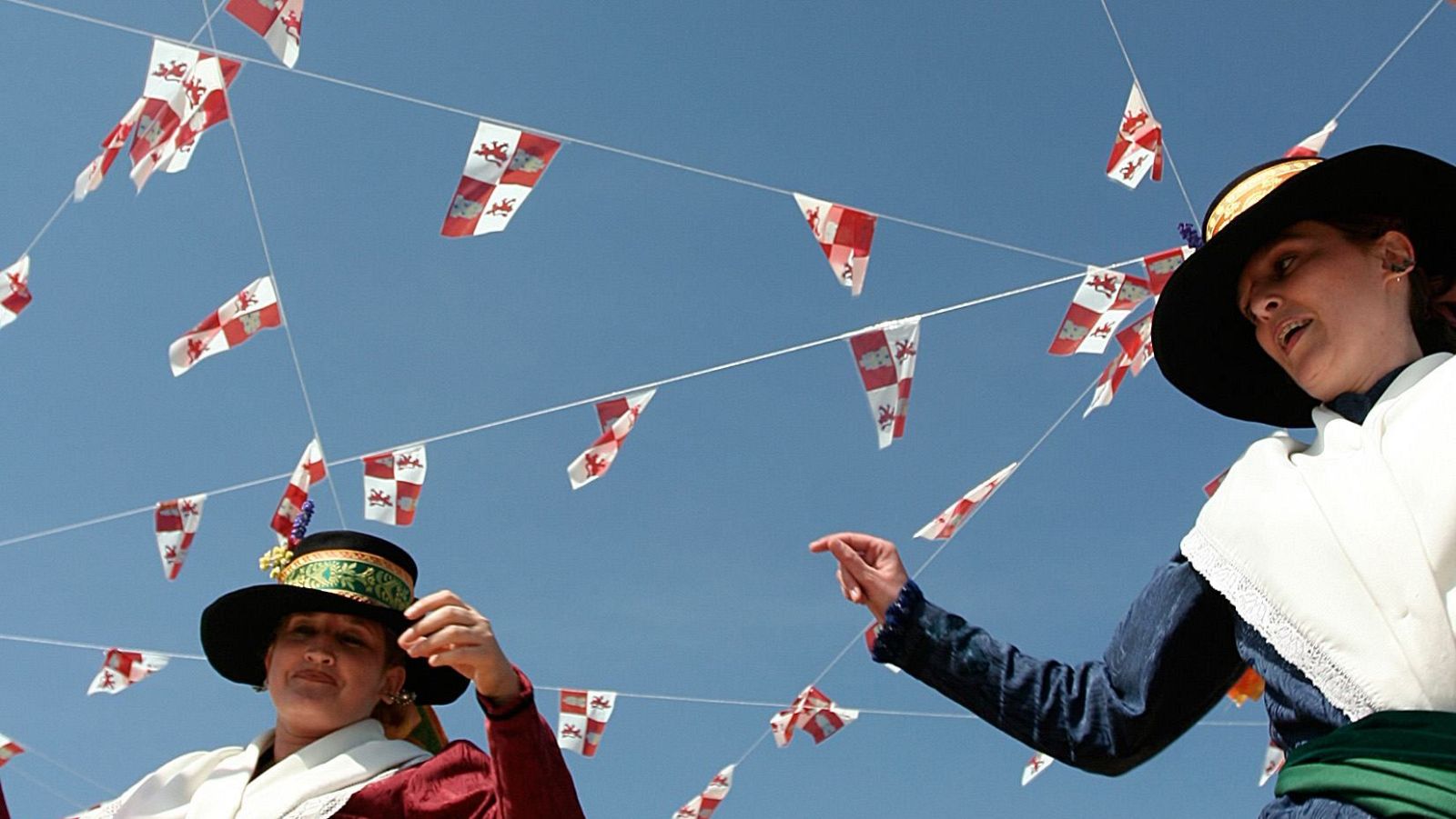 Dos mujeres, ataviadas con trajes tradicionales, bailan bajo un cielo azul adornado con banderas rojas y blancas. Una de ellas lleva un sombrero negro y detalles azules, y la otra, un sombrero con adornos verdes y dorados y detalles rojos.