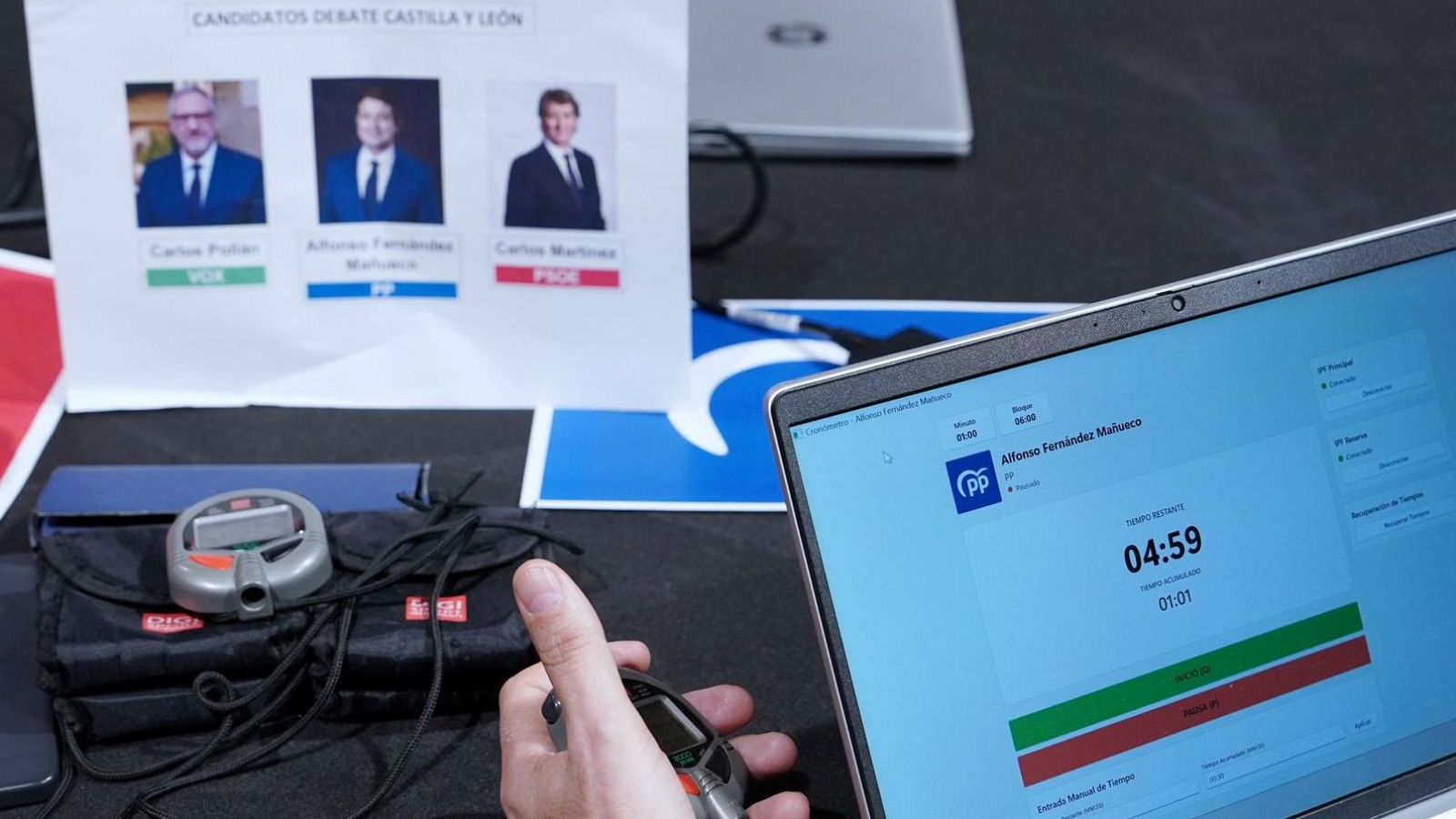 En la sala de control, un ordenador portátil muestra el tiempo restante para un candidato, mientras que un cronómetro manual y un cartel con fotografías de los candidatos completan la escena.