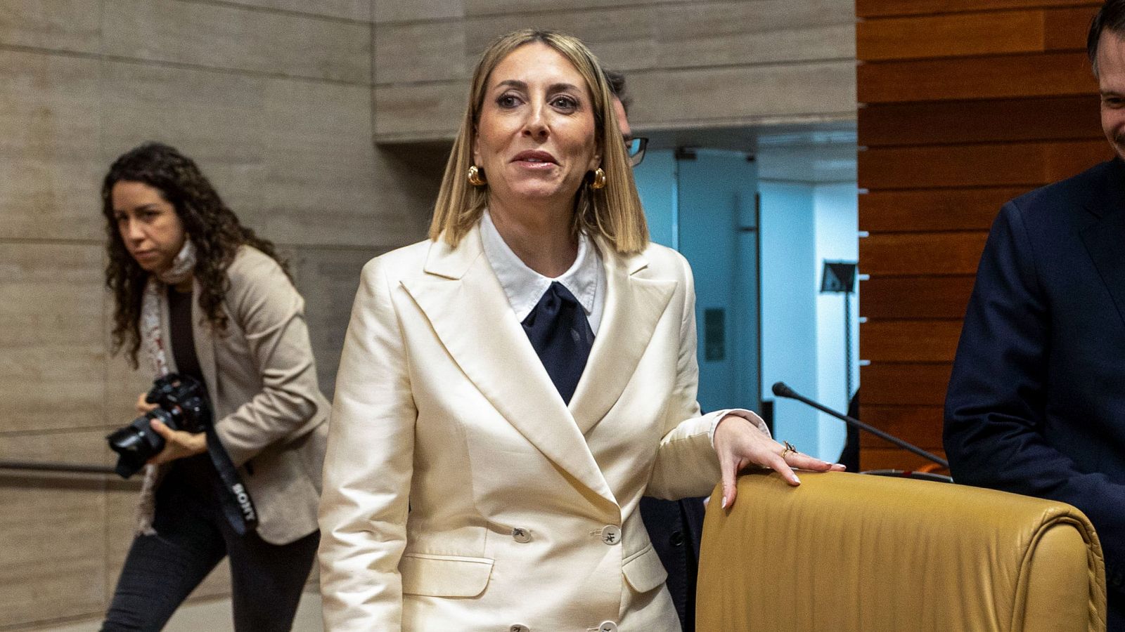 María Guardiola, con un traje blanco y corbata azul, posa en un entorno oficial. Se observa a otras personas en el fondo, incluyendo a una mujer con una cámara fotográfica.