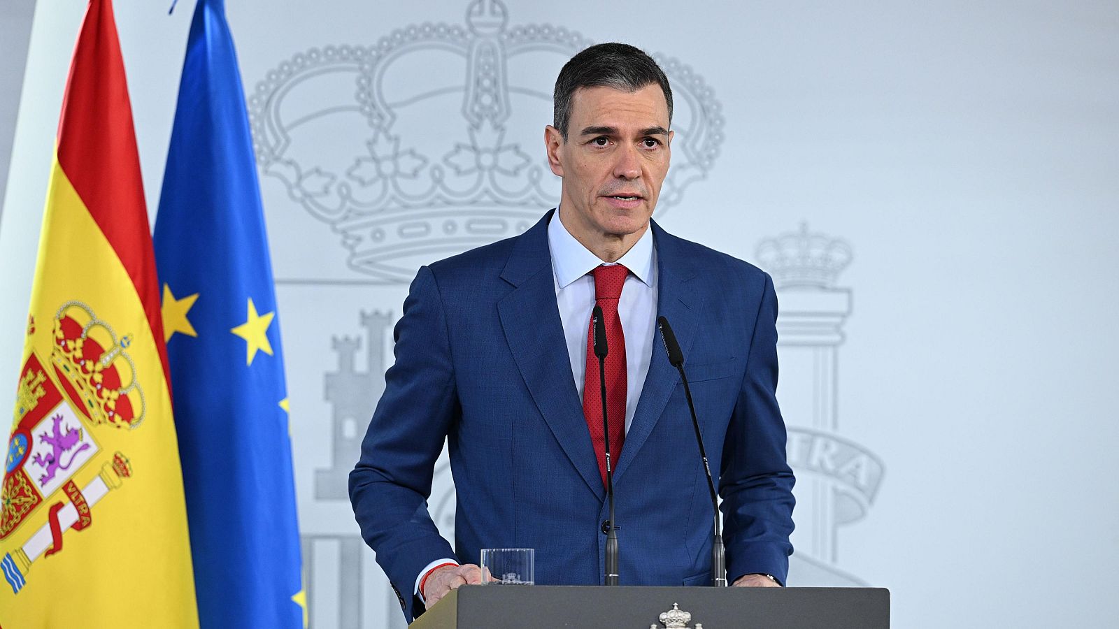 Pedro Sánchez, Presidente del Gobierno, realiza una declaración institucional frente a un atril, vestido con traje azul y corbata roja, con banderas y un escudo al fondo.