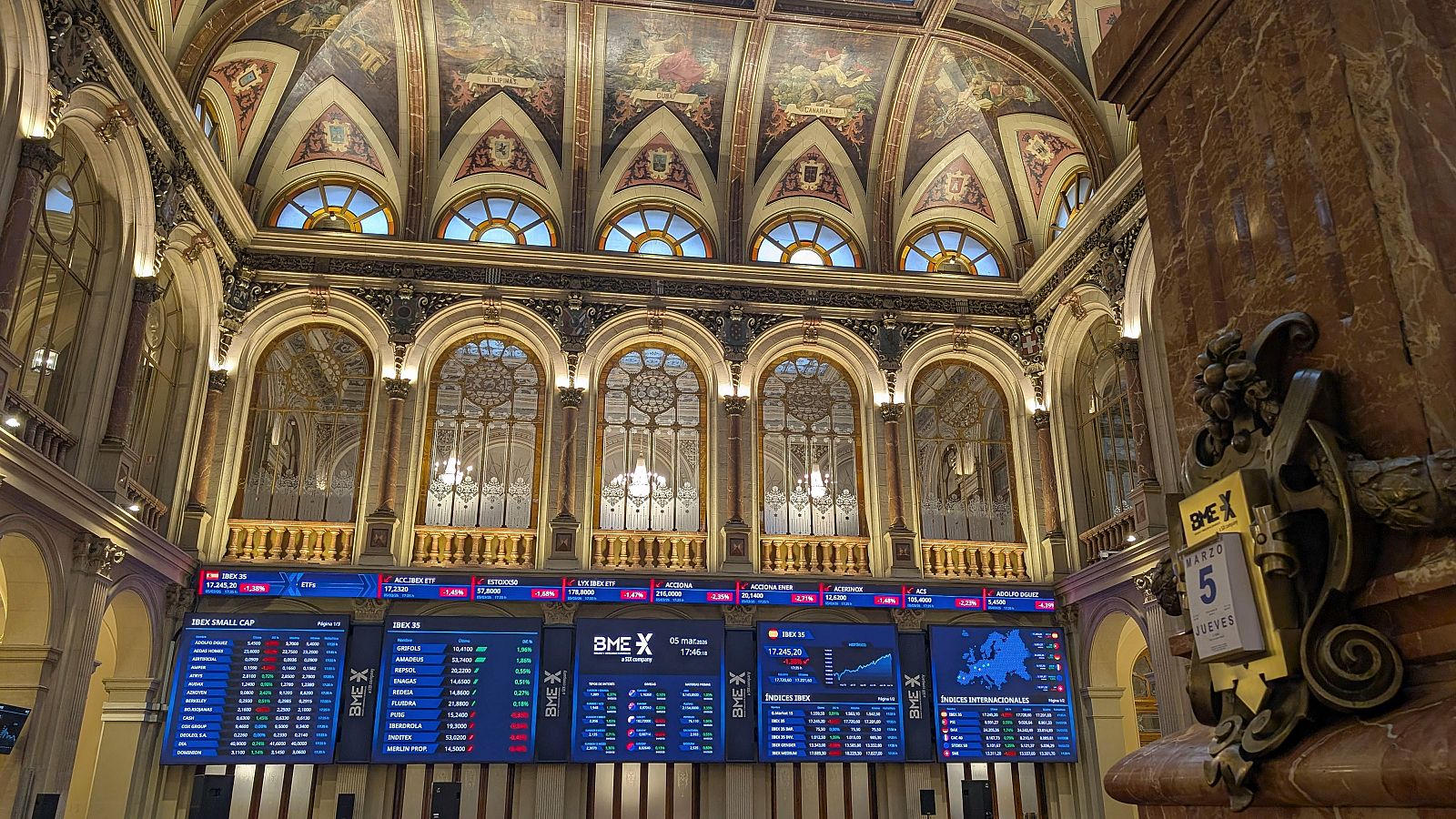Interior de edificio con arquitectura clásica y ornamentada, con paneles electrónicos que muestran información bursátil, incluyendo datos del Ibex 35 y otros índices, y un calendario con la fecha "Marzo 5, Jueves".