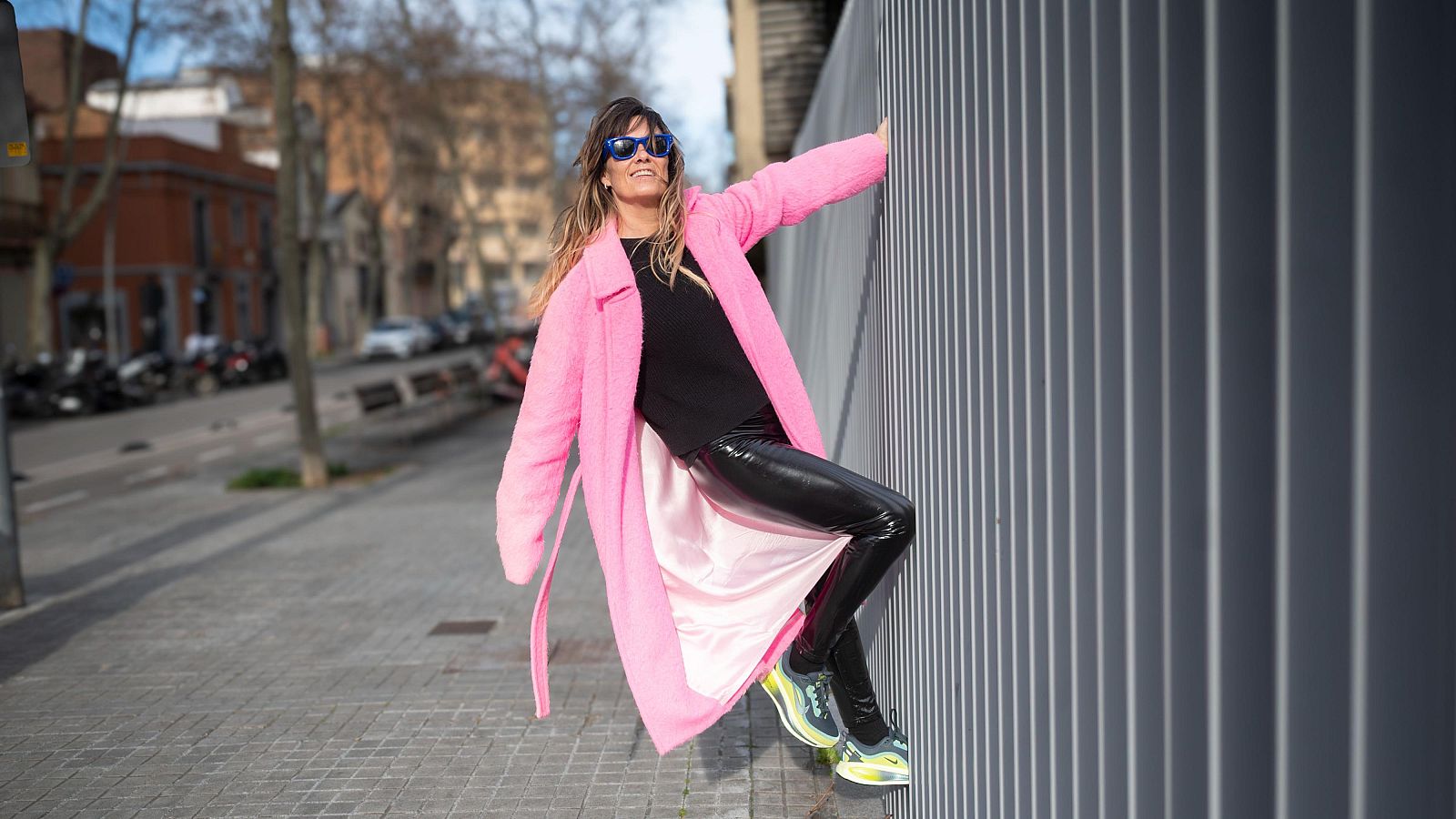 Una periodista amb ulleres de sol i abric rosa brillant posa en un carrer. Vesteix roba fosca i sabatilles esportives, recolzada en una paret grisa amb llistons verticals.