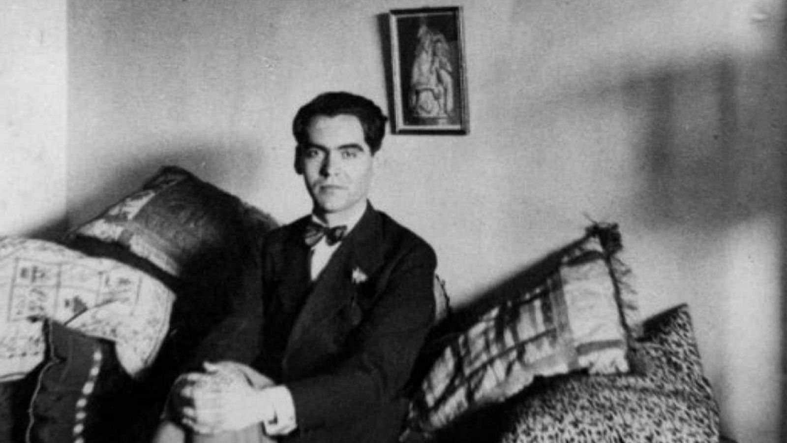 Retrato en blanco y negro de Federico García Lorca, sentado, con traje oscuro, pajarita y expresión seria. Fondo con un cuadro y sofá con cojines.