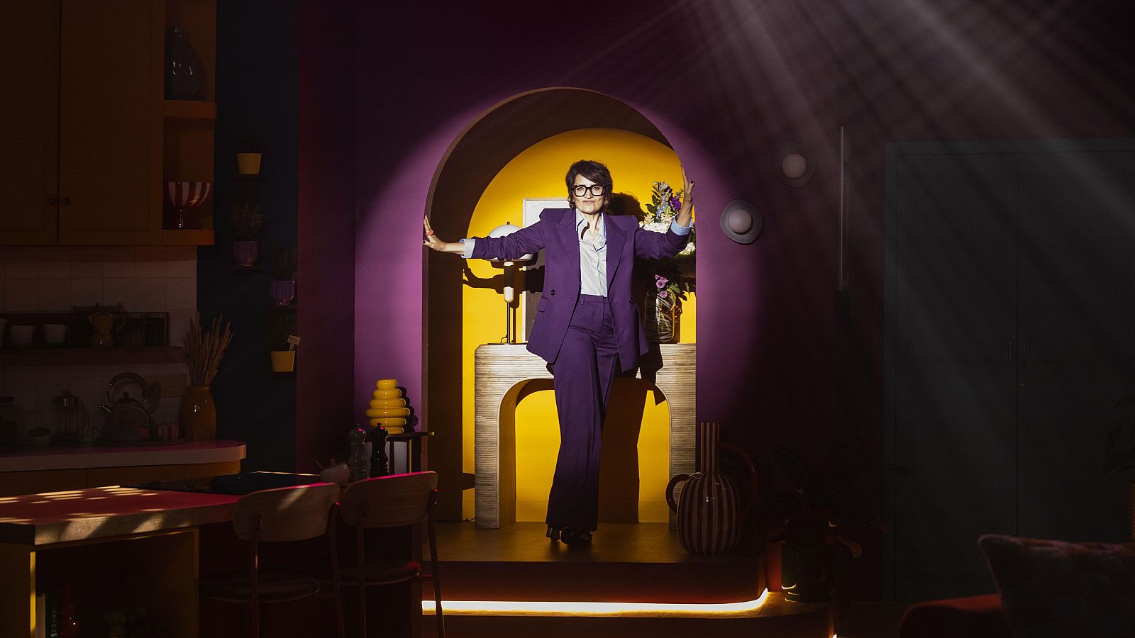 En un plató, Sílvia Abril, vestida amb un conjunt morat, posa davant d'un arc. L'escenografia inclou una habitació groga i una cuina, amb una il·luminació dramàtica que crea ombres marcades.