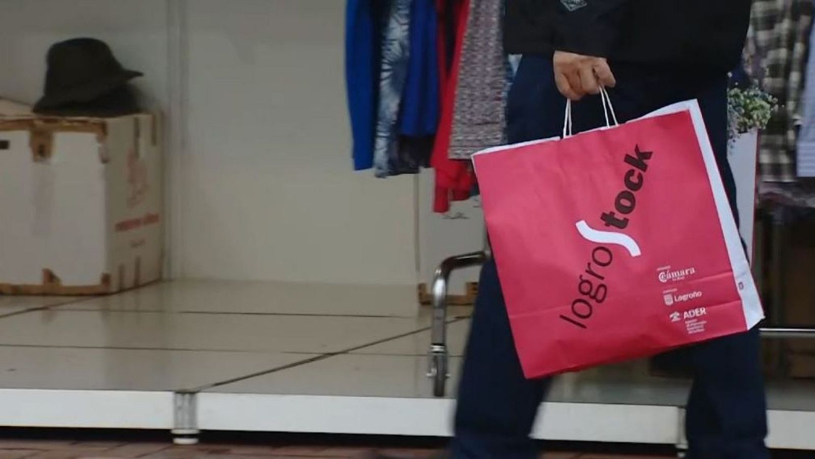 Una bolsa de compras roja con el texto "logroStock" y los logos de "Cámara Logroño" y "ADER" es el foco principal de la imagen, con ropa y un sombrero en el fondo.