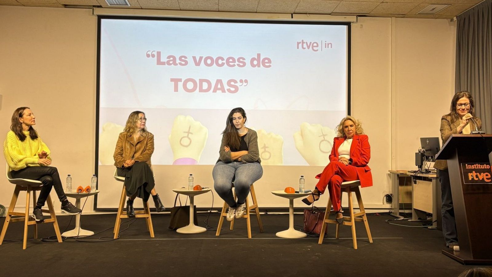 Un panel de discusión con cinco mujeres, cada una con un estilo de vestimenta distinto, se presenta frente a una pantalla con el texto "Las voces de TODAS" y el logo de una entidad.