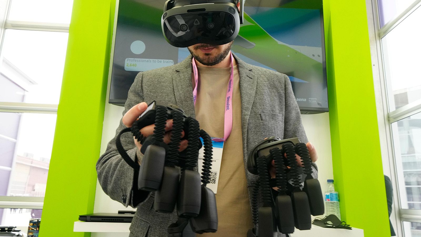 En un stand, un hombre con barba y chaqueta gris utiliza gafas de realidad virtual y guantes hápticos. Se observa una cinta rosa con un gafete colgando de su cuello, y una pantalla con información en el fondo.