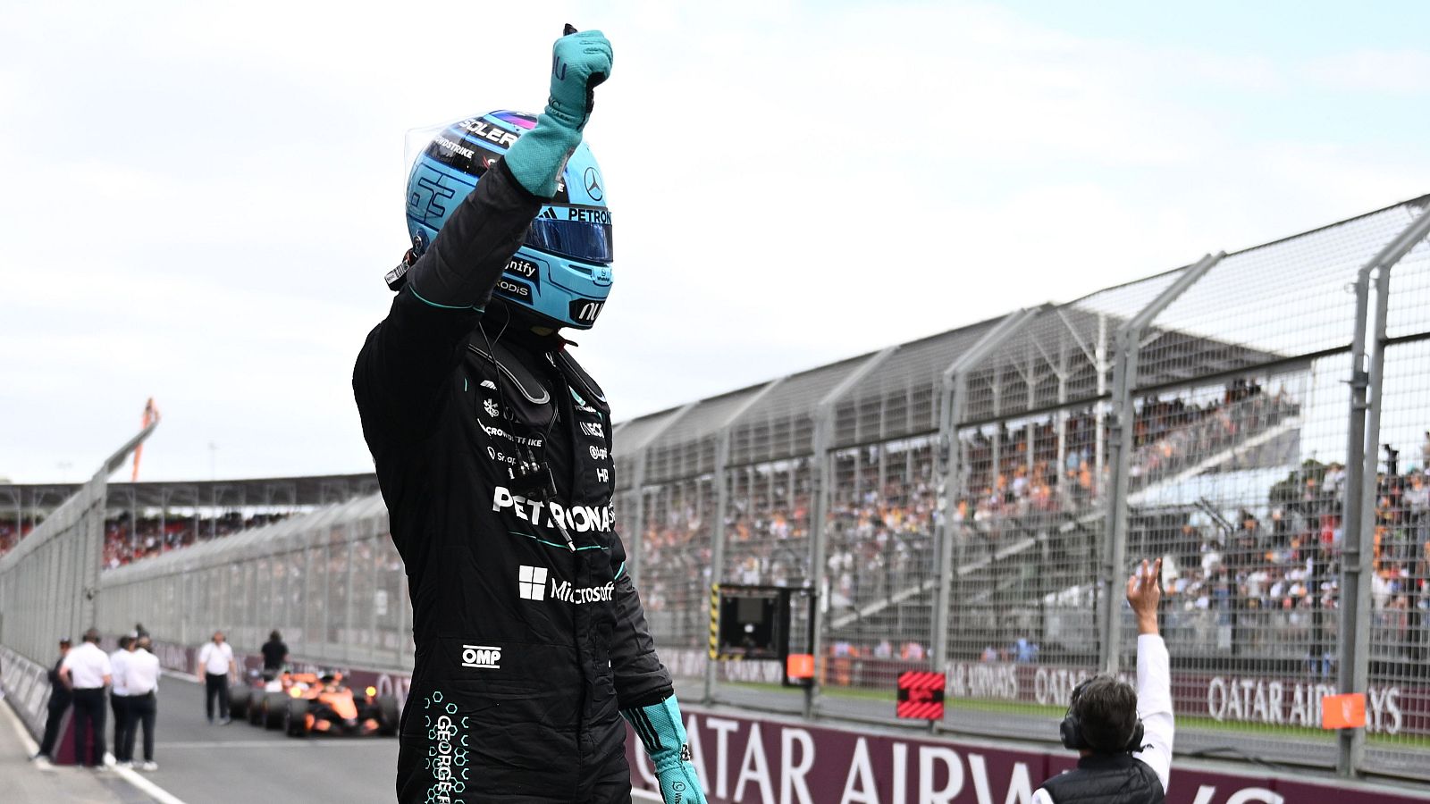 George Russell, con su mono de carreras y casco, celebra la pole position en un circuito, levantando el brazo en señal de victoria. Se observan gradas llenas de público y una valla publicitaria.