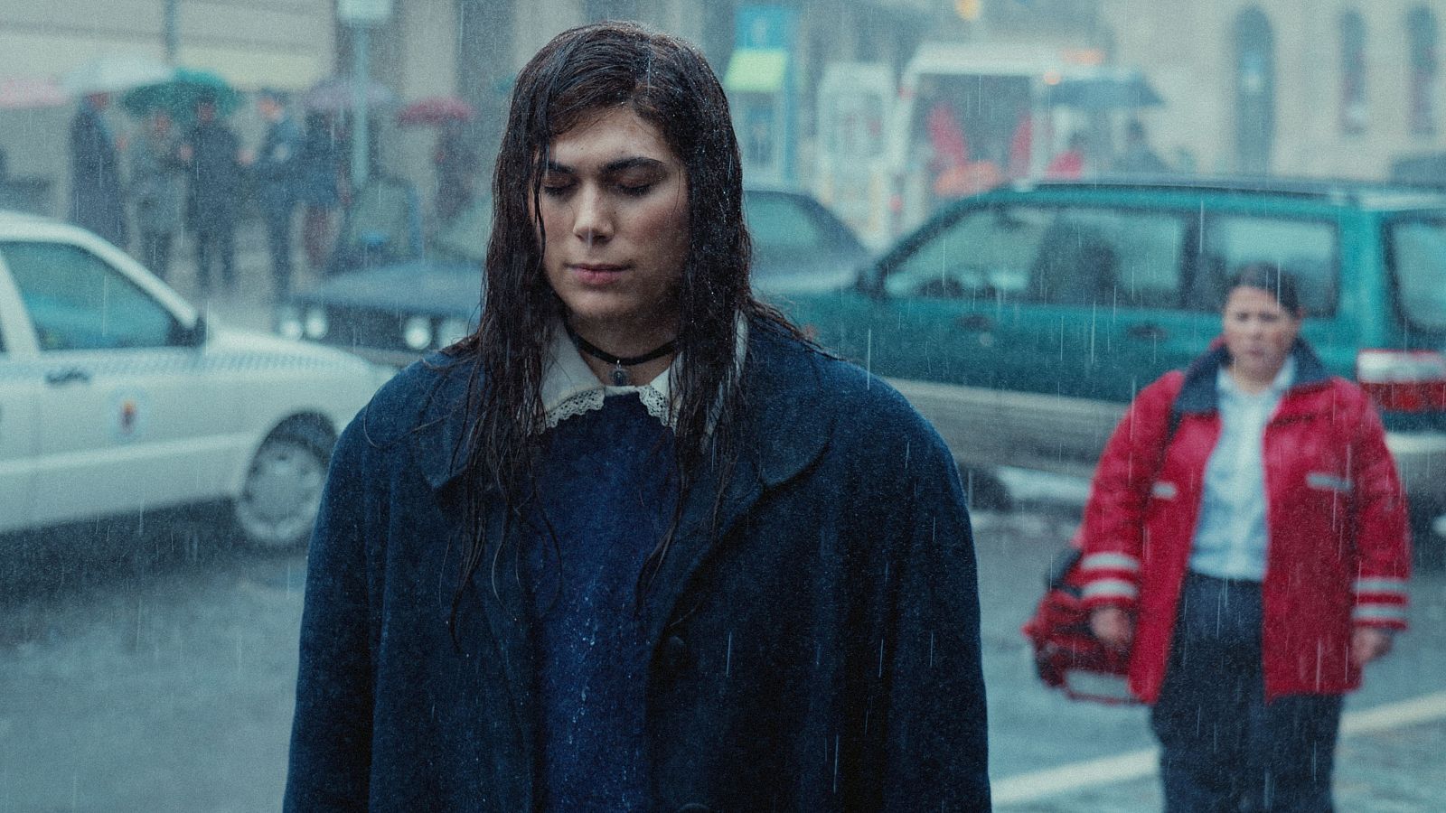 Una joven con vestido azul oscuro y abrigo, de mirada melancólica, se encuentra en una calle lluviosa con coches y peatones, en un fotograma de una película.