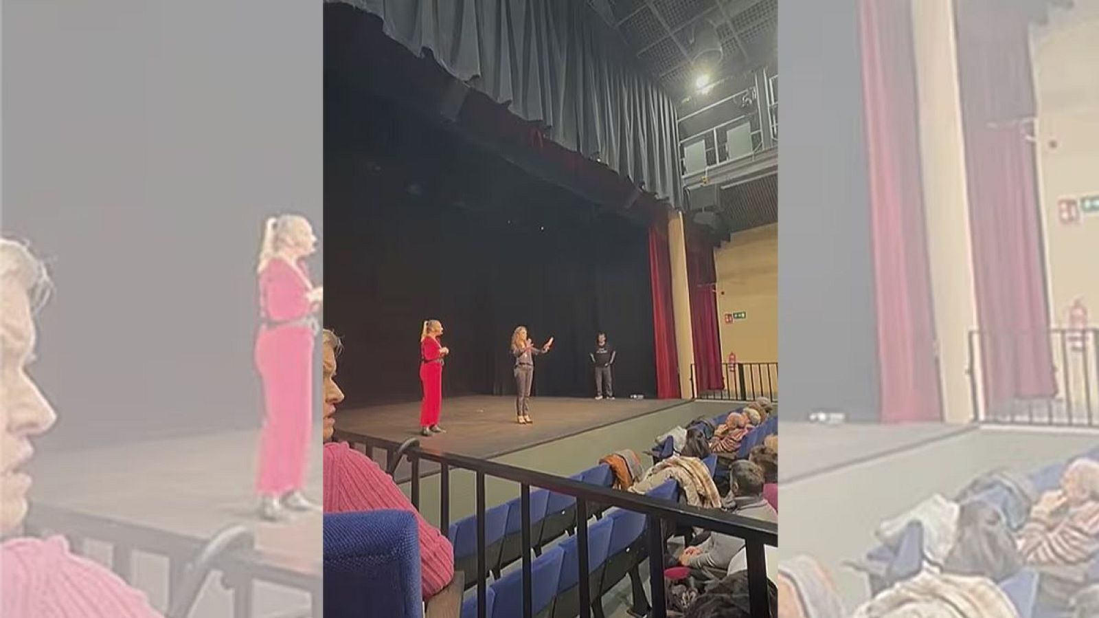 En un teatro iluminado, tres figuras se destacan en el escenario: una mujer de rojo, otra de gris con micrófono y un hombre de negro. El público, diverso, ocupa las butacas frente al escenario.