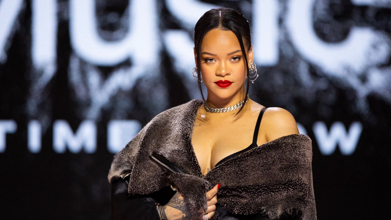 Imagen de Rihanna con chaqueta oscura, pelo recogido y labios rojos. Se ven tatuajes en una mano y parte del texto "MUSIC TIME NOW" en el fondo.