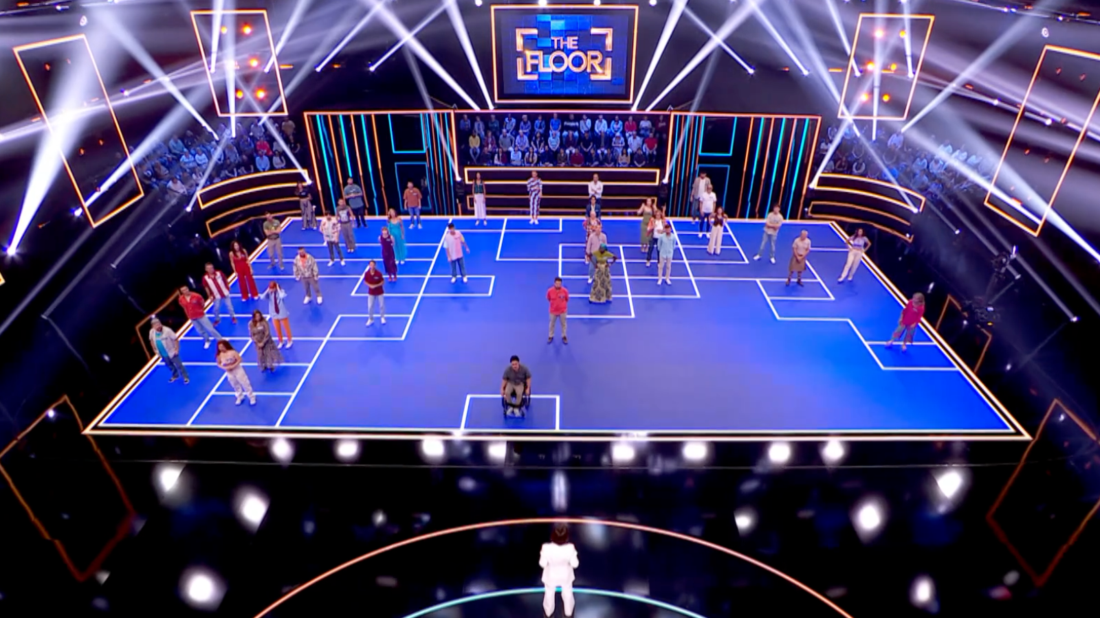 En un escenario azul con cuadrados blancos, concursantes de "The Floor" compiten, incluyendo a uno en silla de ruedas. El presentador, de espaldas y vestido de blanco, observa la dinámica del juego.