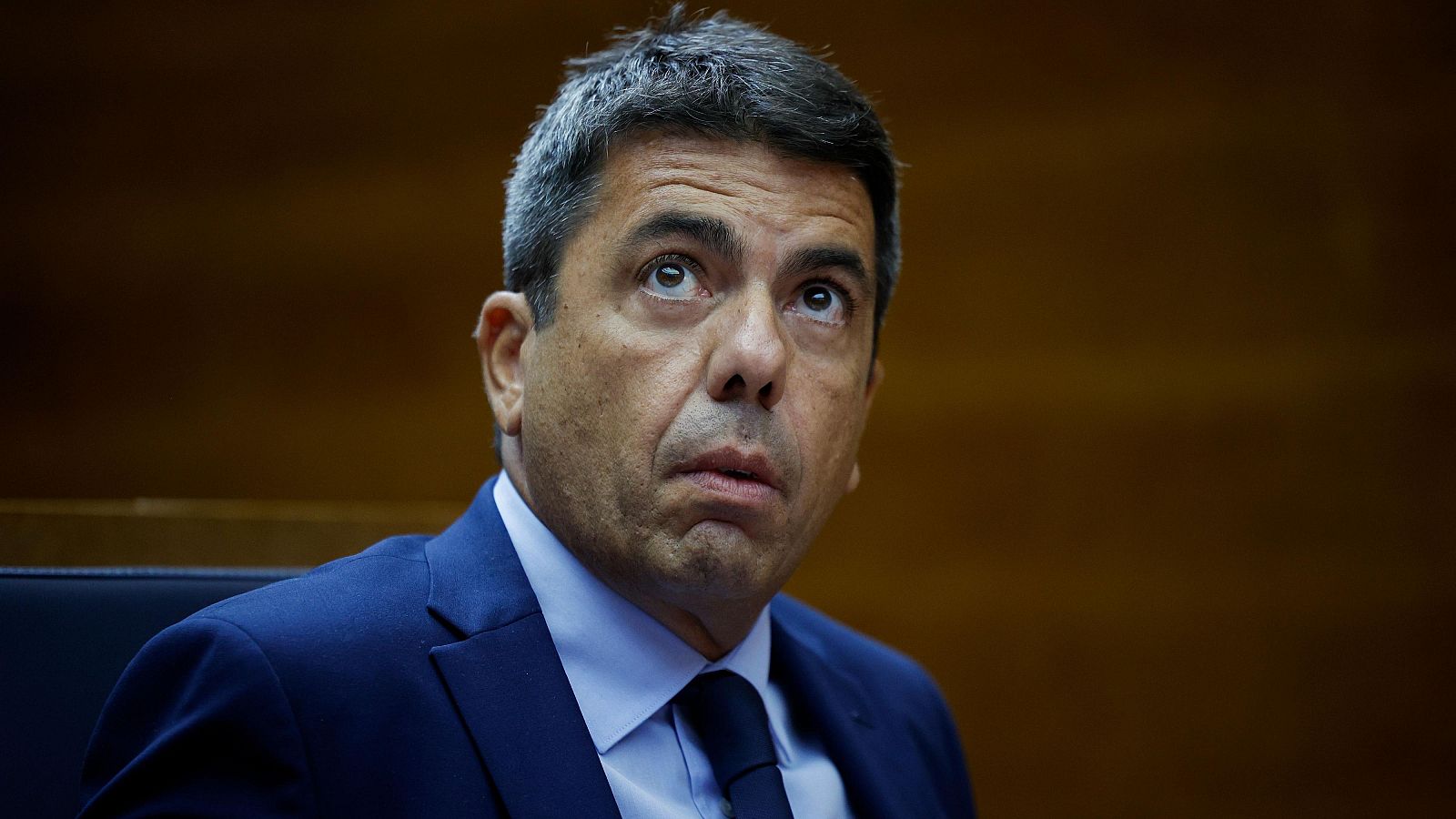 Primer plano de un hombre con traje oscuro, camisa blanca y corbata, mirando hacia arriba. El fondo es una pared de madera.
